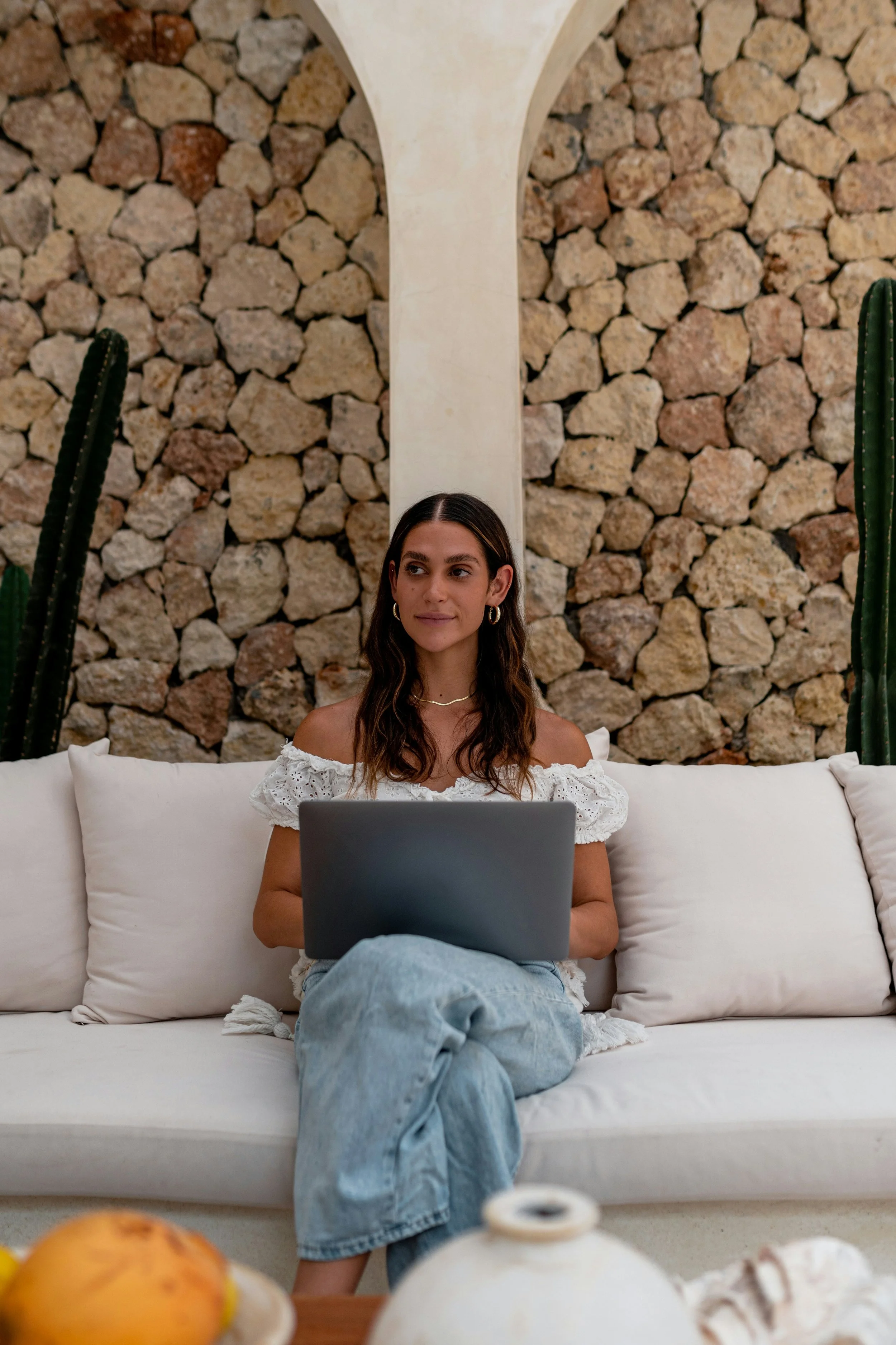 A young woman sitting on a white couch with a laptop on her lap, in front of a stone wall. She has long brown hair, wears a white off-shoulder top and light blue jeans. There are tall cacti on either side of her.