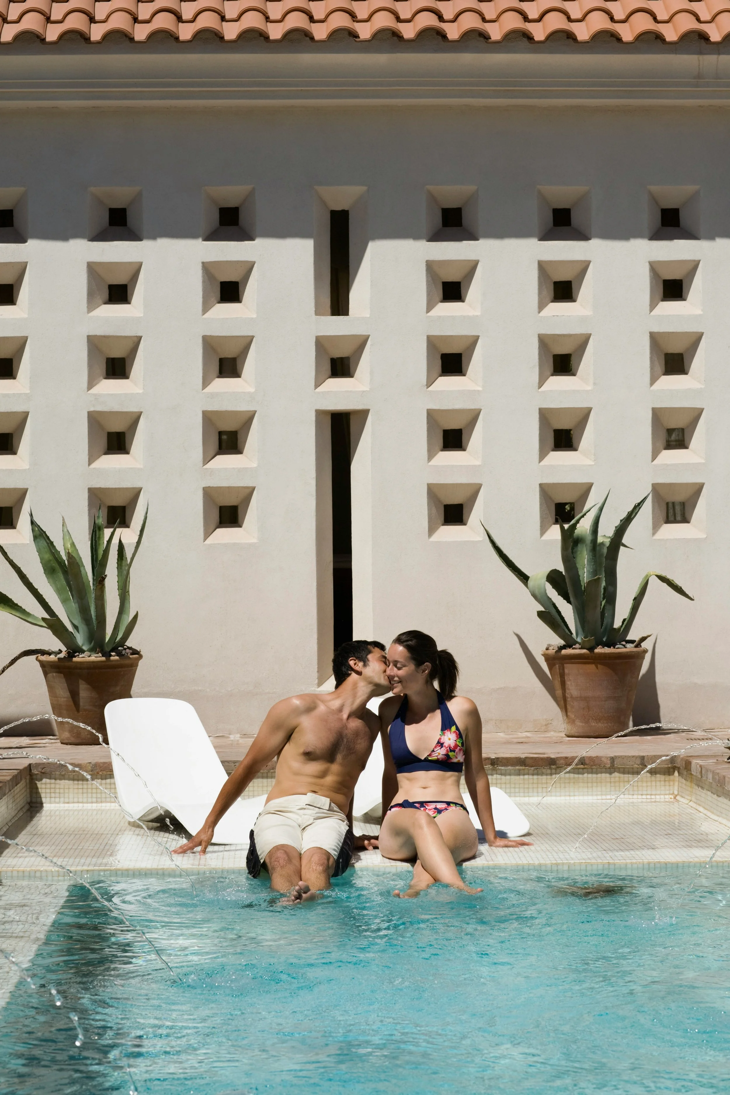 A man and woman sit on the edge of a swimming pool, sharing a kiss with their legs in the water, surrounded by potted agave plants and a decorative wall.