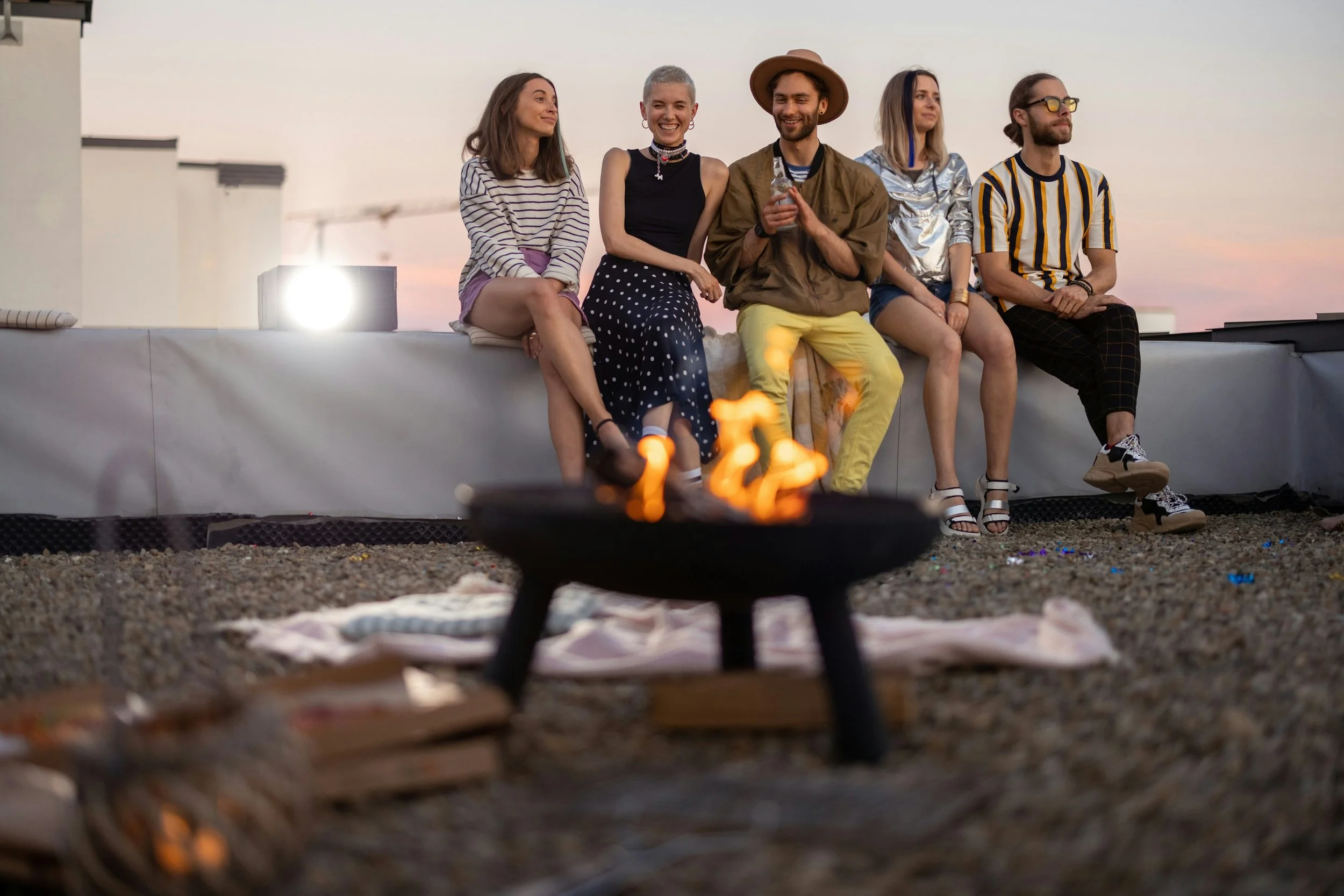 Group of five young adults sitting on a rooftop ledge during sunset, with a small fire pit in the foreground.