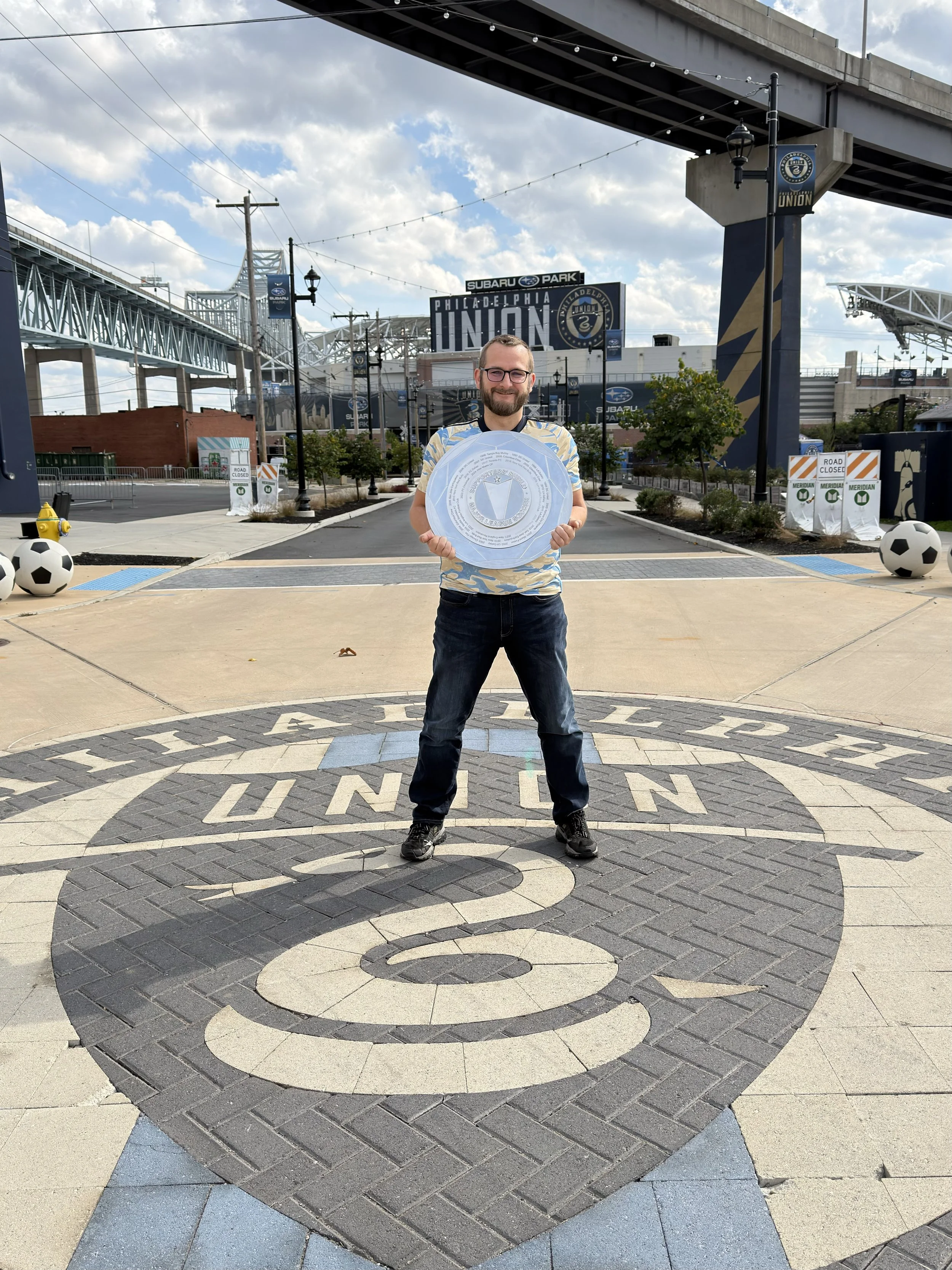 A man standing outdoors at the Philadelphia Union logo on the ground, holding a large circular shield, with the Philadelphia Union stadium in the background.