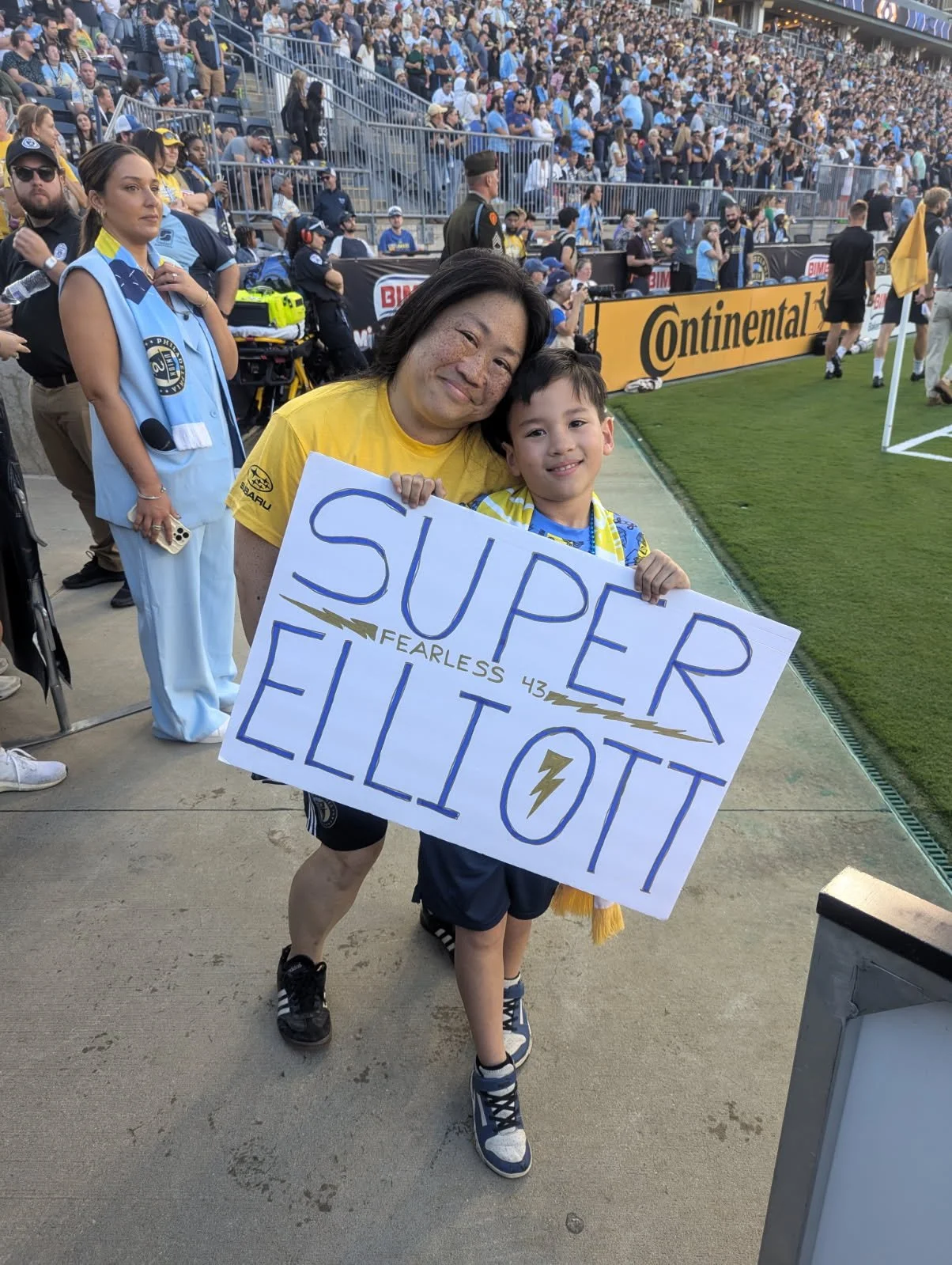 A woman and a young boy on a soccer field holding a sign that reads 'Super Elliott' at a crowded stadium in the background.