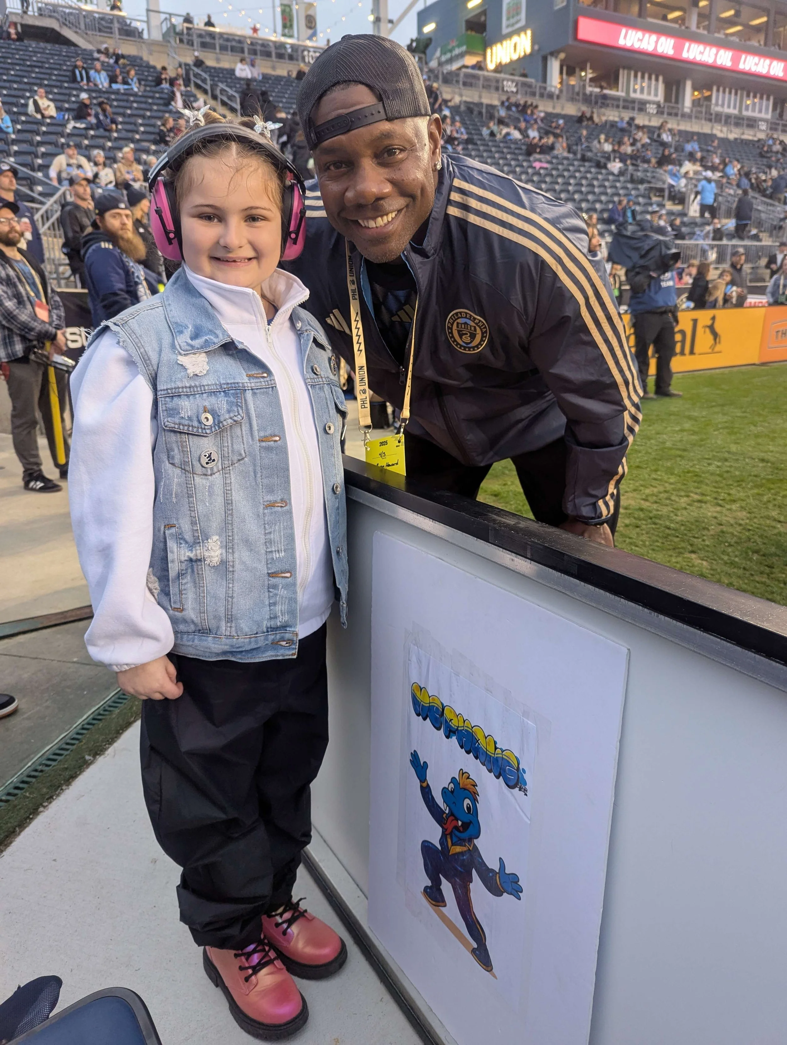 A young girl wearing a denim vest, white hoodie, black pants, pink boots, and pink headphones standing next to a smiling man in a baseball stadium. The man is wearing a black baseball cap, jacket, and a yellow badge. They are near a fence with a cart