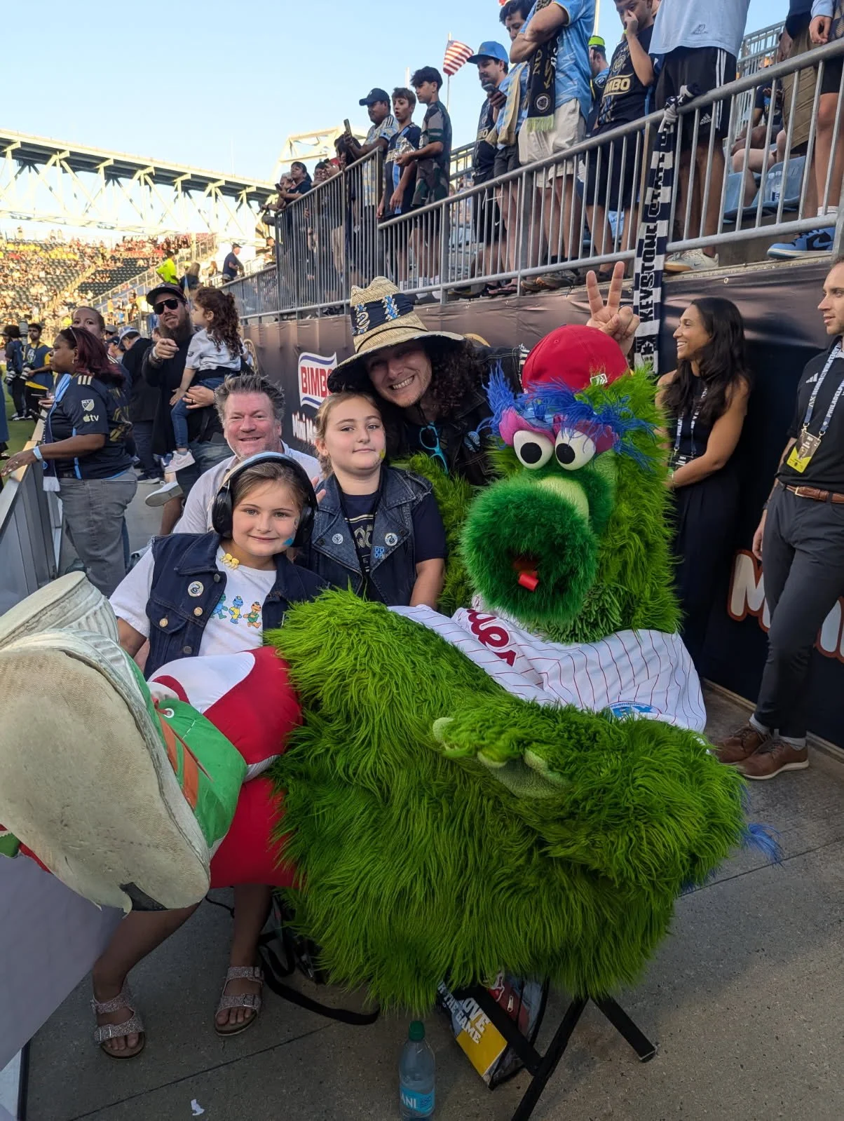A group of children and adults at a sports stadium with a person in a green monster costume. The children are smiling, and the mascot is sitting on a table. The background shows more fans and a stadium seating area.