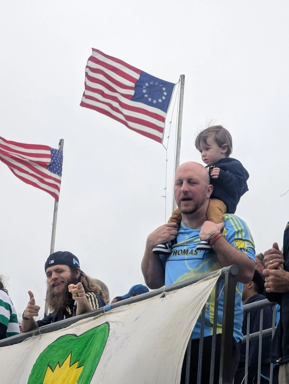 A group of people at an outdoor event, with two American flags and one South Carolina state flag flying in the background. A man with a child on his shoulders, and another man giving a thumbs-up and smiling.