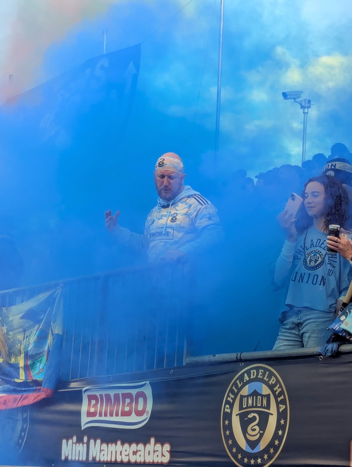 People standing behind a barrier at a sports event, some wearing Philadelphia Union jerseys, surrounded by blue smoke or fog.