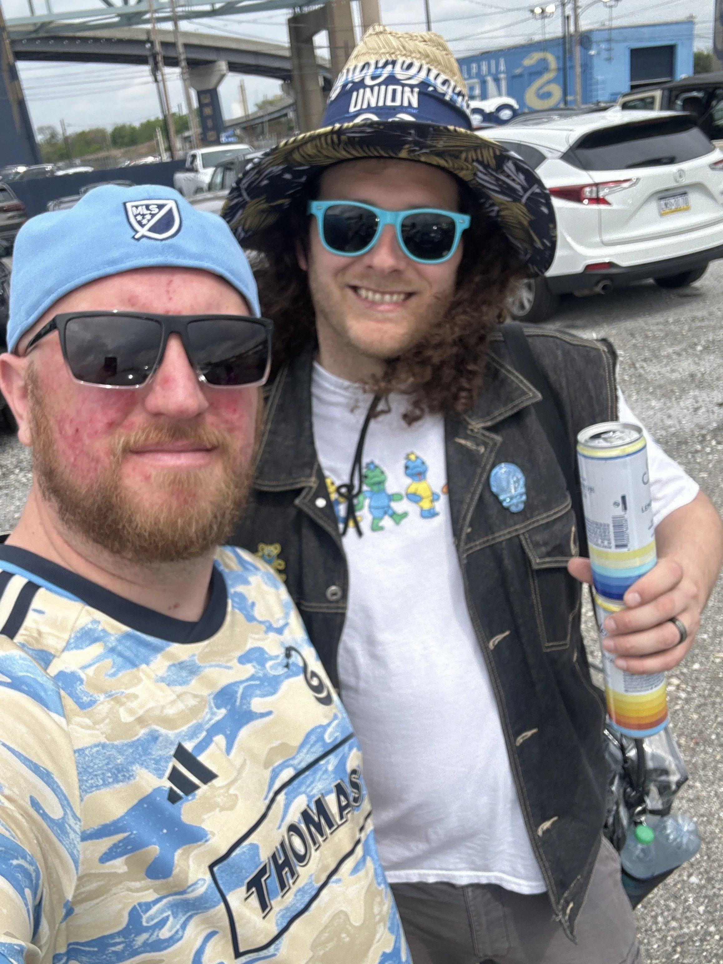 Two men taking a selfie outdoors in a parking lot. The man in front wears a light blue hat, sunglasses, and a patterned shirt. The man behind wears a wide-brimmed hat, large blue sunglasses, a denim vest, and holds a can of drink.