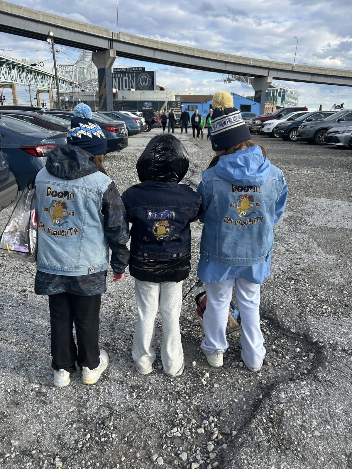 Three people wearing denim jackets with the logo 'Boom DOLLIMANTS' on their backs, standing in a parking lot under a bridge, with other cars and people in the background, some wearing beanies and warm clothing.
