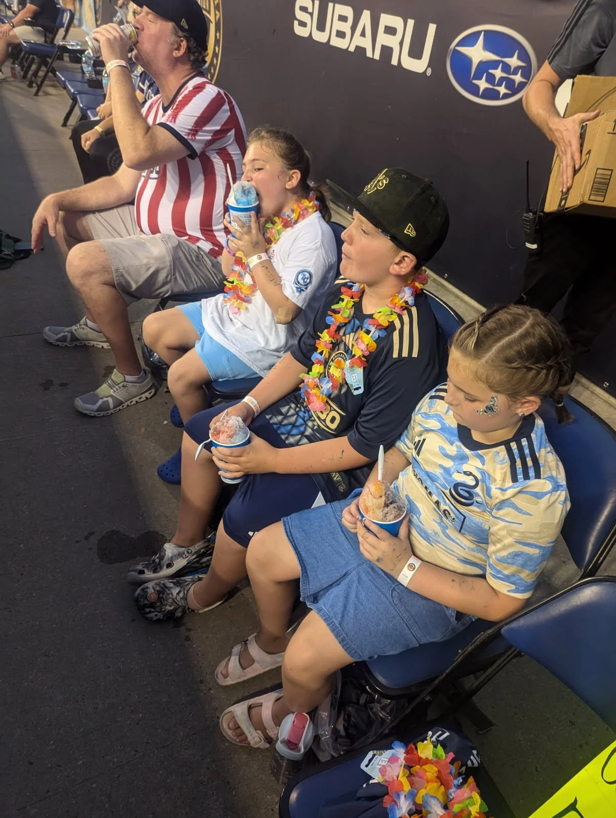 Three children and a man sitting on chairs, enjoying snow cones at an event. The children are wearing leis, and some are dressed in sports jerseys. A man in a striped shirt and swimming trunks is drinking, with a large Subaru sign in the background.