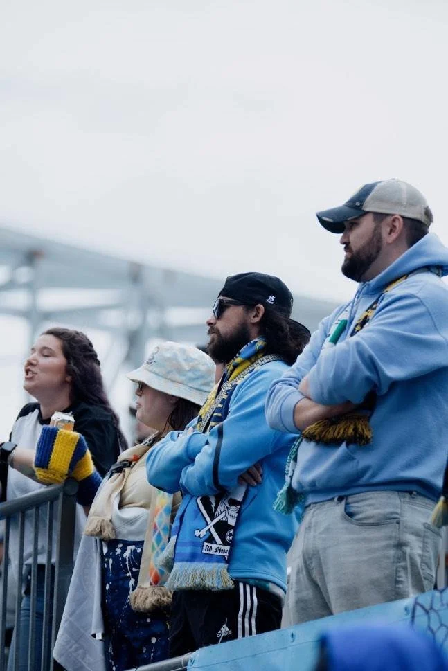 Group of sports fans standing behind a barrier, watching a game outdoors on a cloudy day. They are wearing team apparel and scarves.