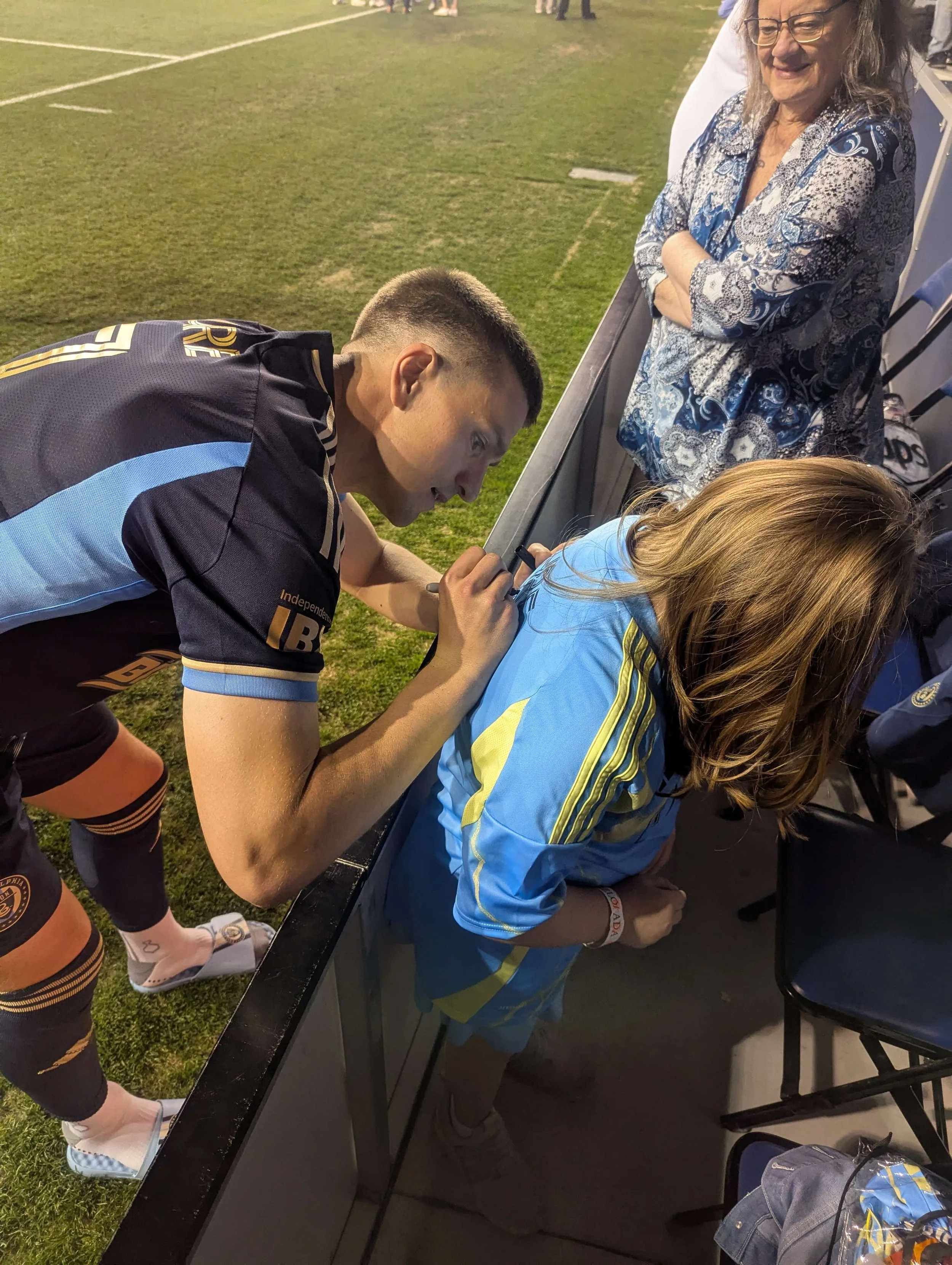 A male football player in a black and blue uniform signing an autograph on a female fan's blue and yellow sports jersey at a stadium.