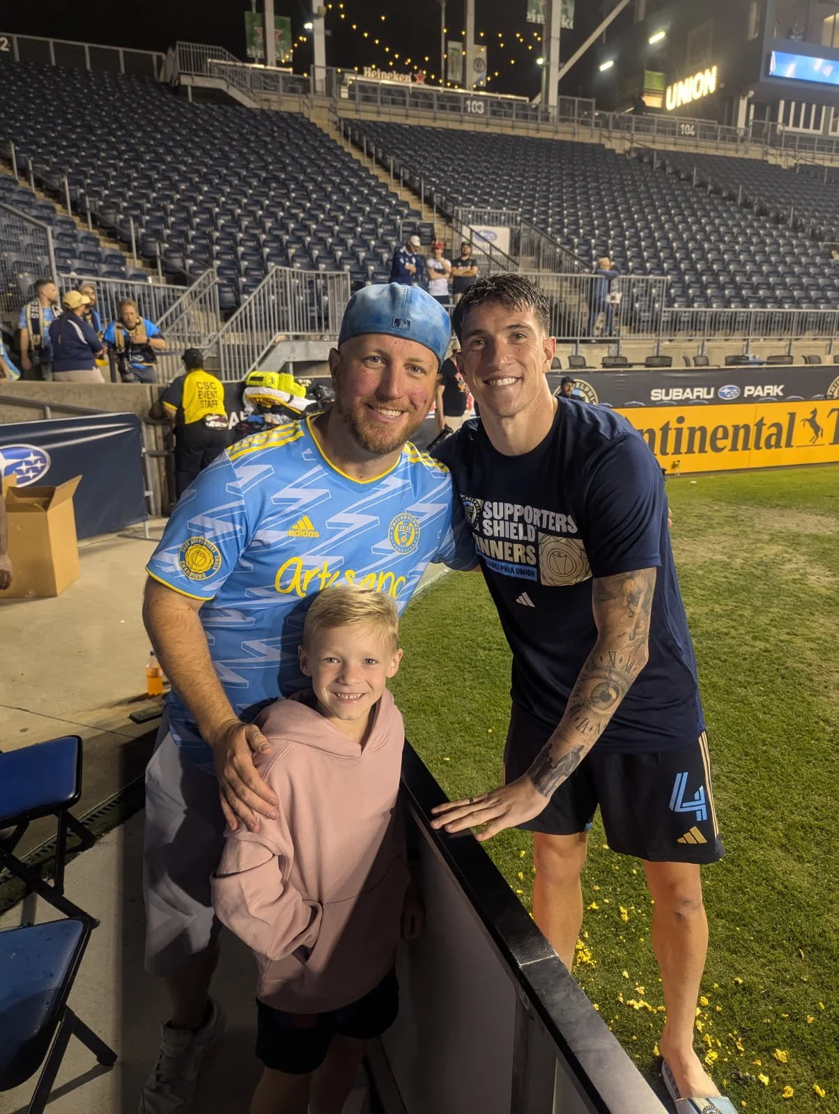 Three people, two men and a child, posing for a photo at a sports stadium. The two men are standing on either side of the child, all smiling. The man on the left is wearing a light blue sports jersey and a blue cap, the man on the right is wearing a 
