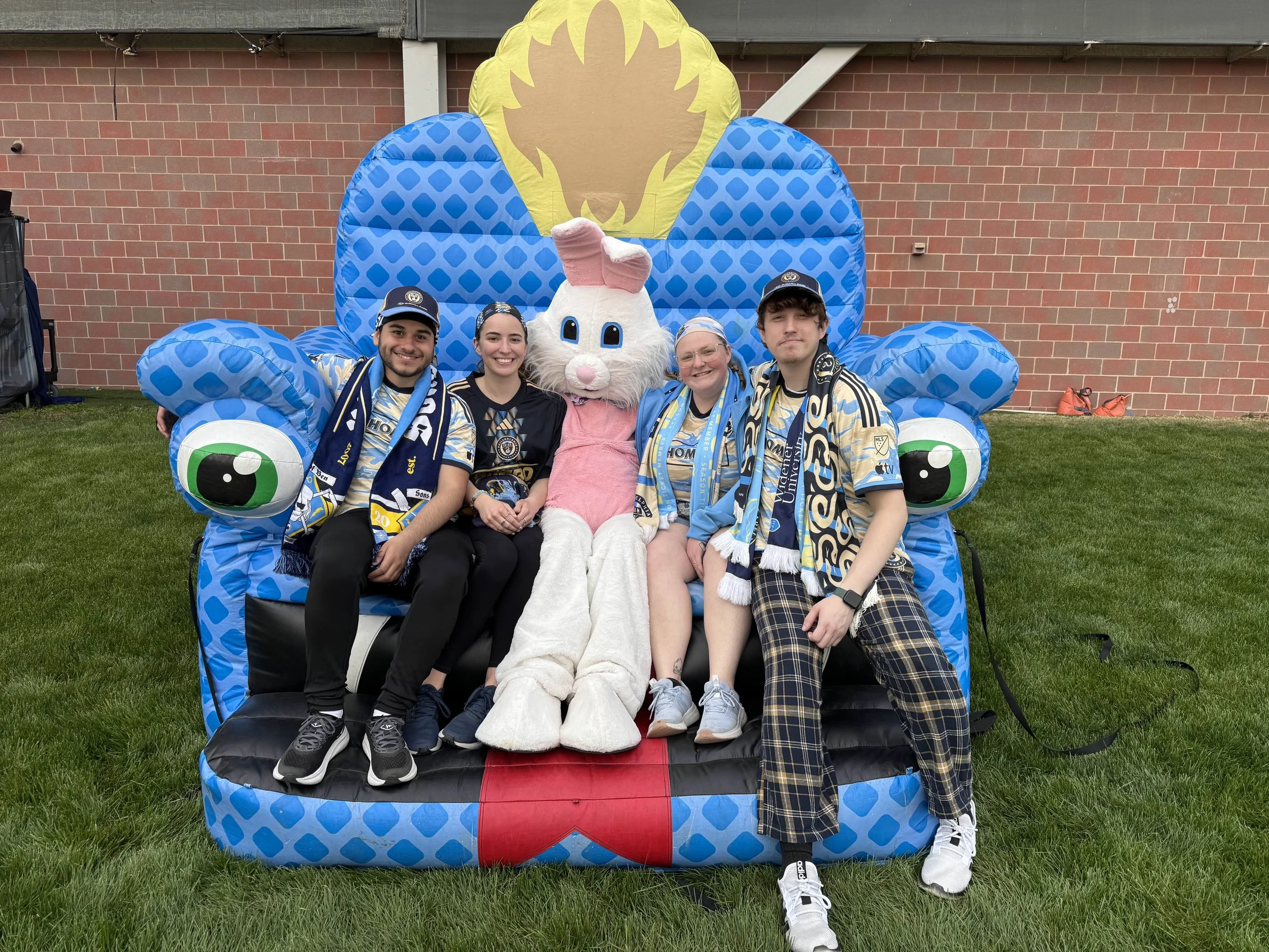 Four people sitting on a large, colorful inflatable seat with a bunny mascot in the center, all wearing matching sports team scarves and hats, outdoors on grass with a brick wall in the background.