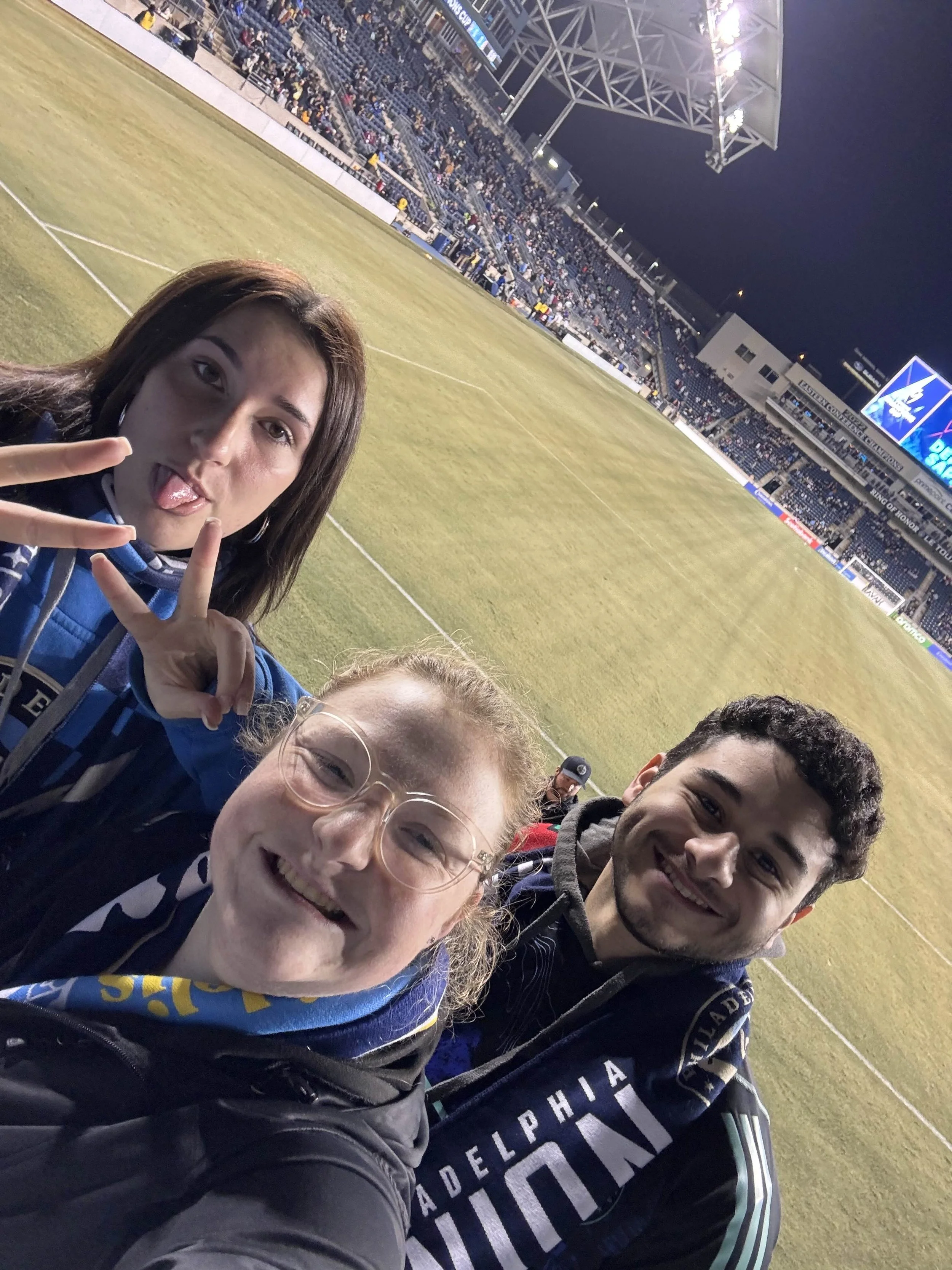 Three friends taking a selfie at a sports stadium during a game, with the field and audience in the background.