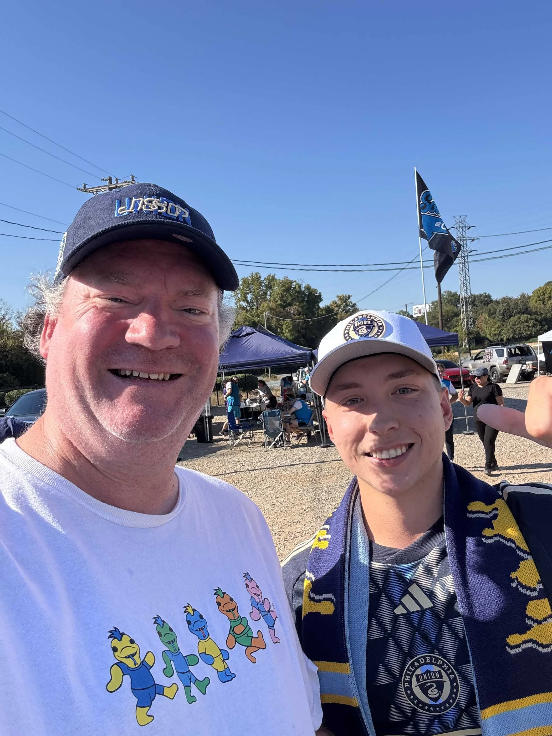 Two smiling men at an outdoor event, wearing sports caps and casual clothing. The older man is wearing a white T-shirt with a colorful cartoon character print, and the younger man is wearing a sports jersey, a scarf, and a cap with the Philadelphia U