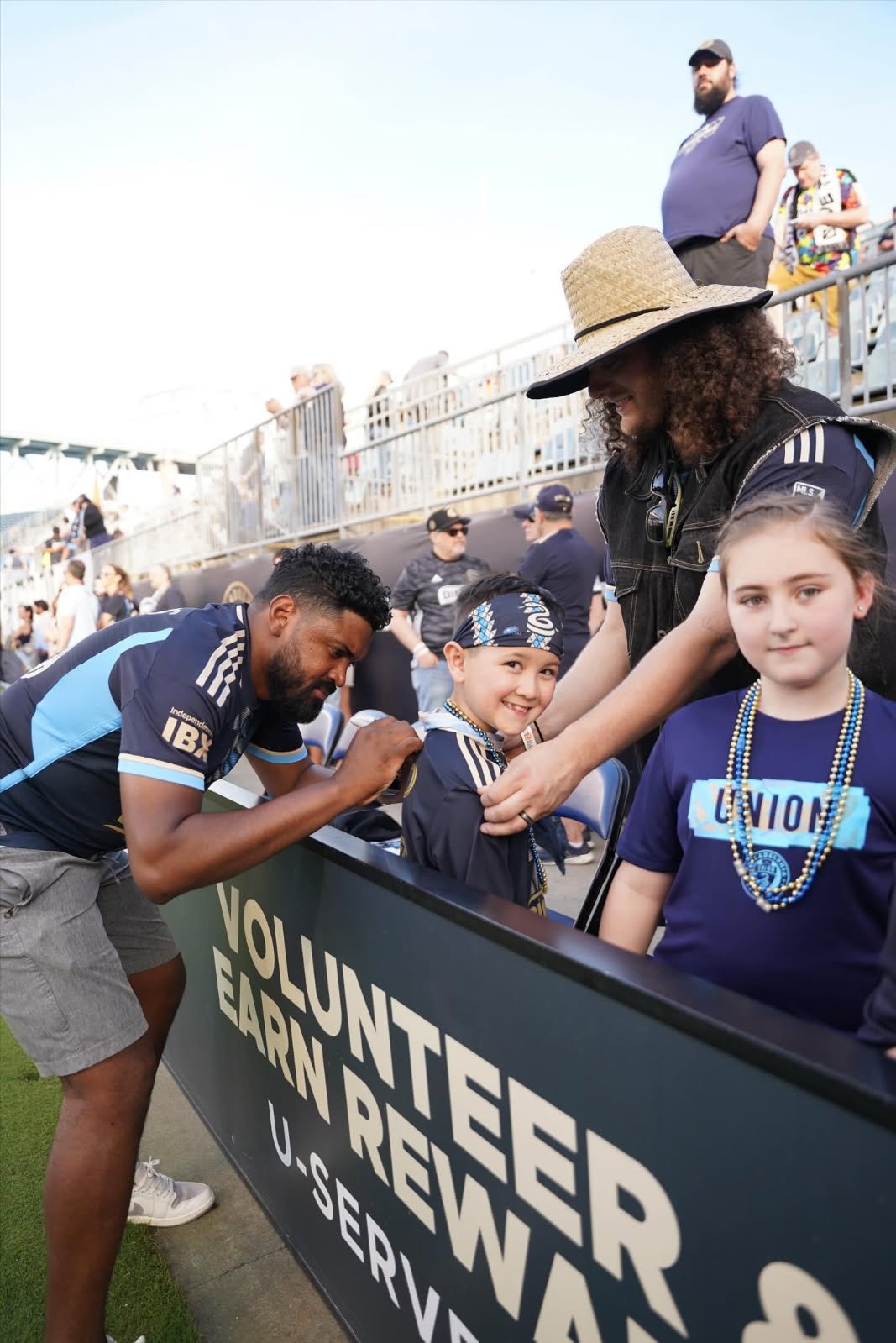Children and adults at a volunteer event, some wearing jerseys and beads, signing jerseys behind a sign that reads "VOLUNTEER, EARN, REWARD, U-SER".