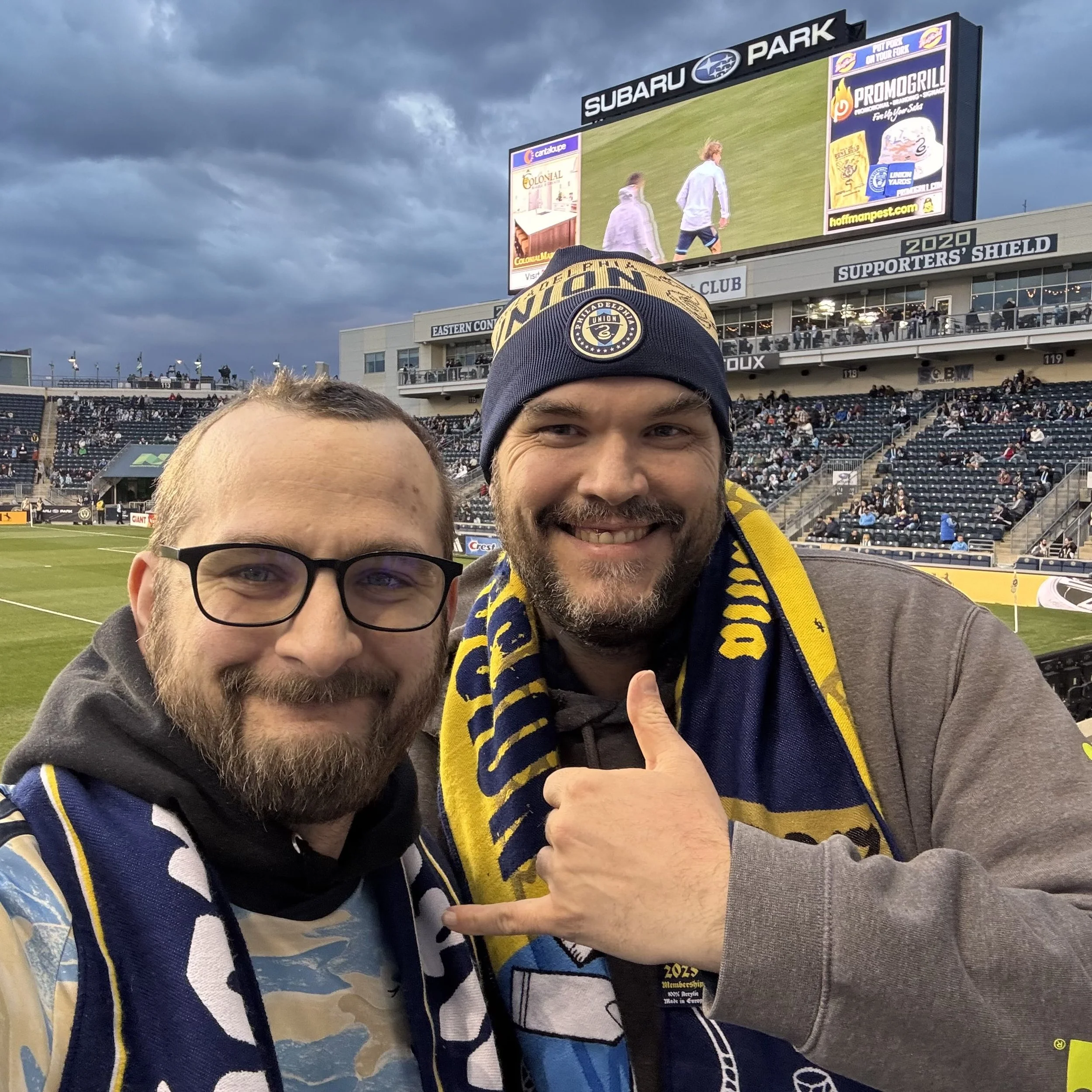 Two men smiling at a soccer game, one wearing glasses and a scarf, and the other wearing a beanie and scarf, with a stadium and a large screen displaying players in the background.