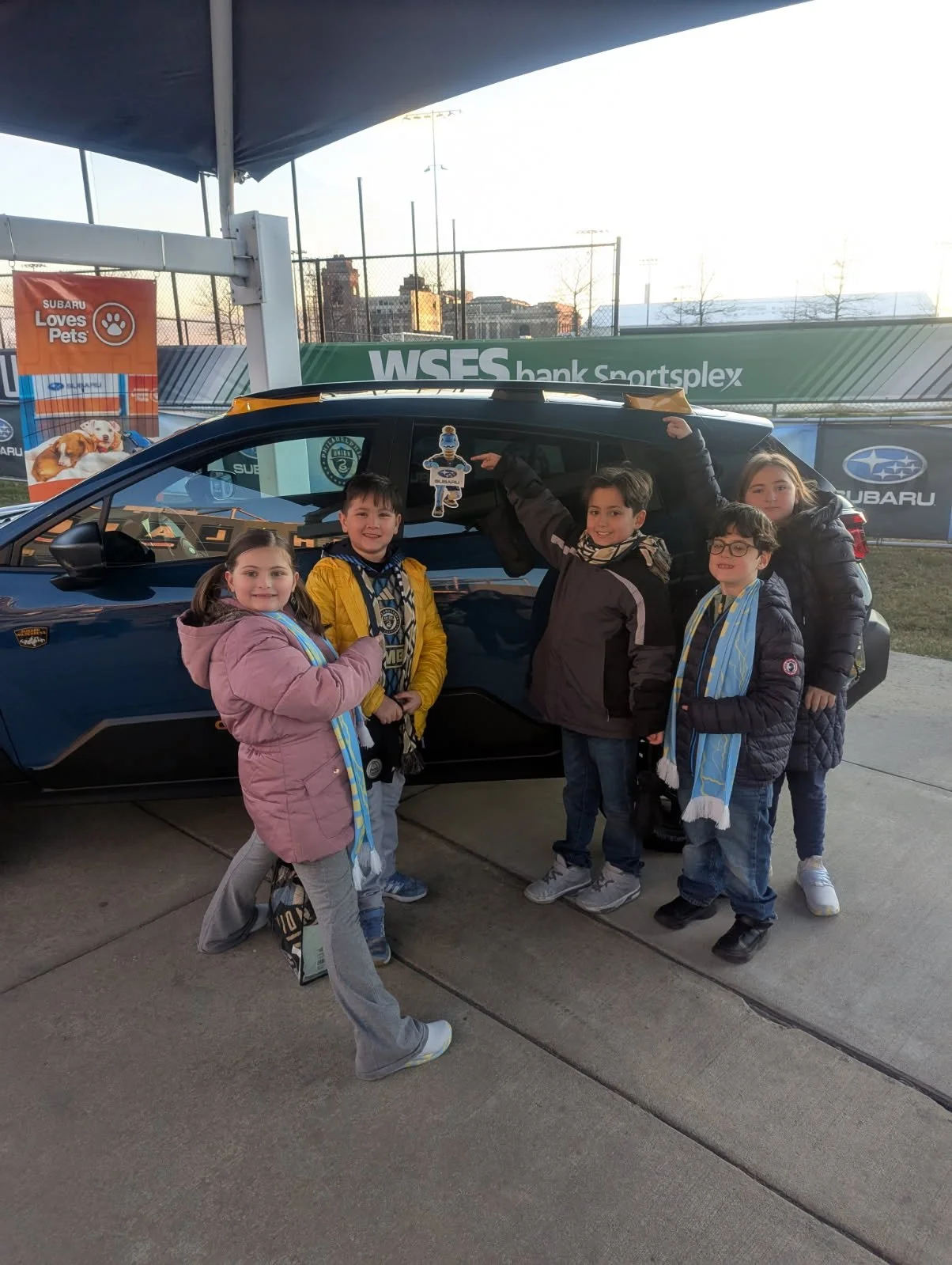 Group of children standing next to a dark blue Subaru car at a sports complex, pointing and smiling, wearing scarves and winter jackets.