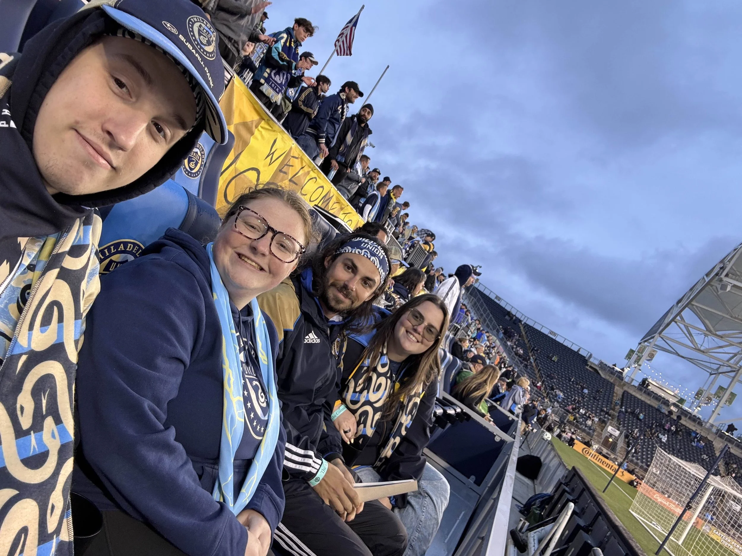 Group of four people sitting in a stadium, wearing team scarves and jackets, with a soccer field and other spectators in the background.