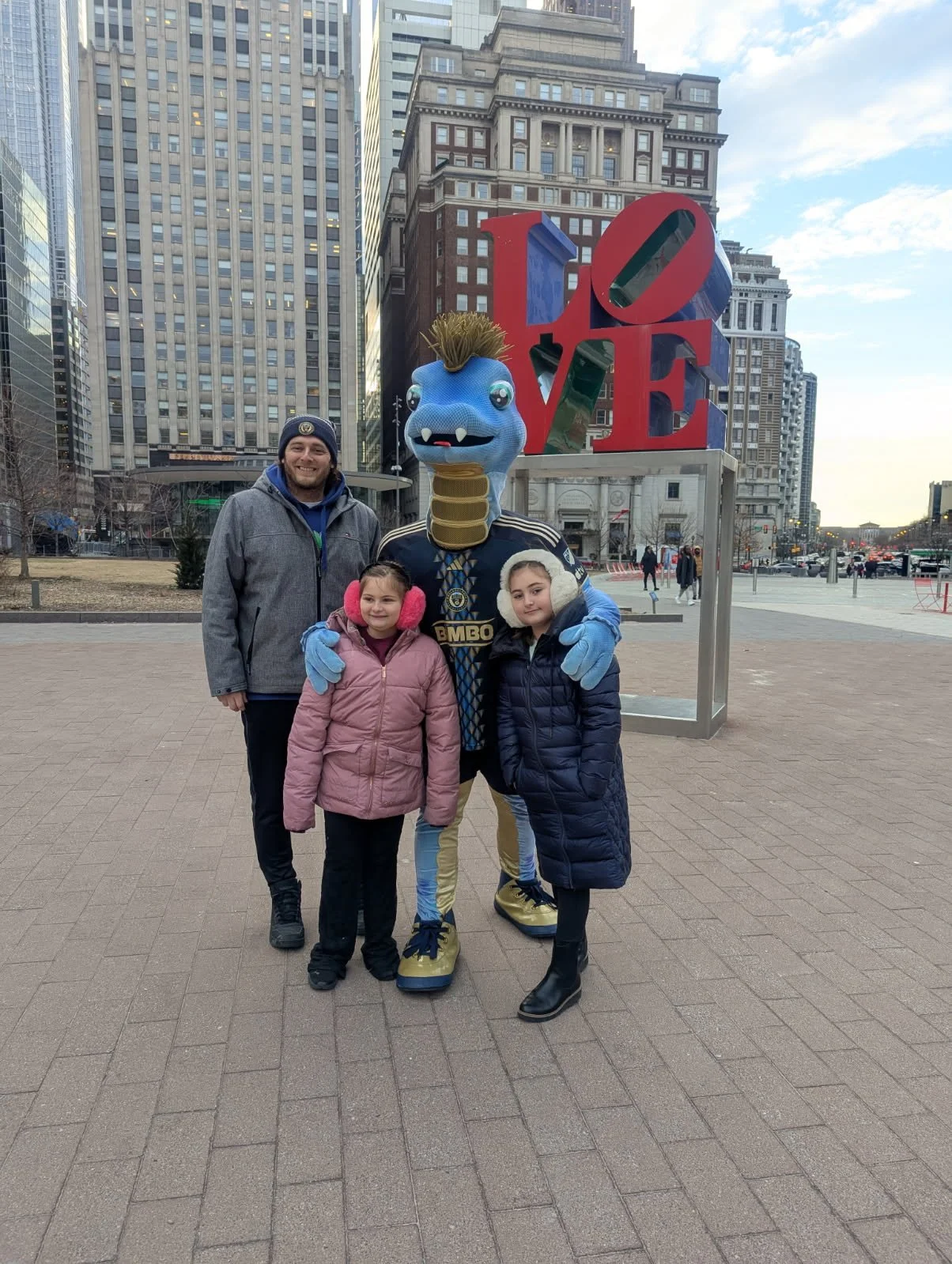 A man and two young girls standing with a mascot at Millennium Park in Chicago, in front of the 'LOVE' sculpture. The mascot has a blue face, large eyes, a mohawk, and is dressed in a soccer jersey.