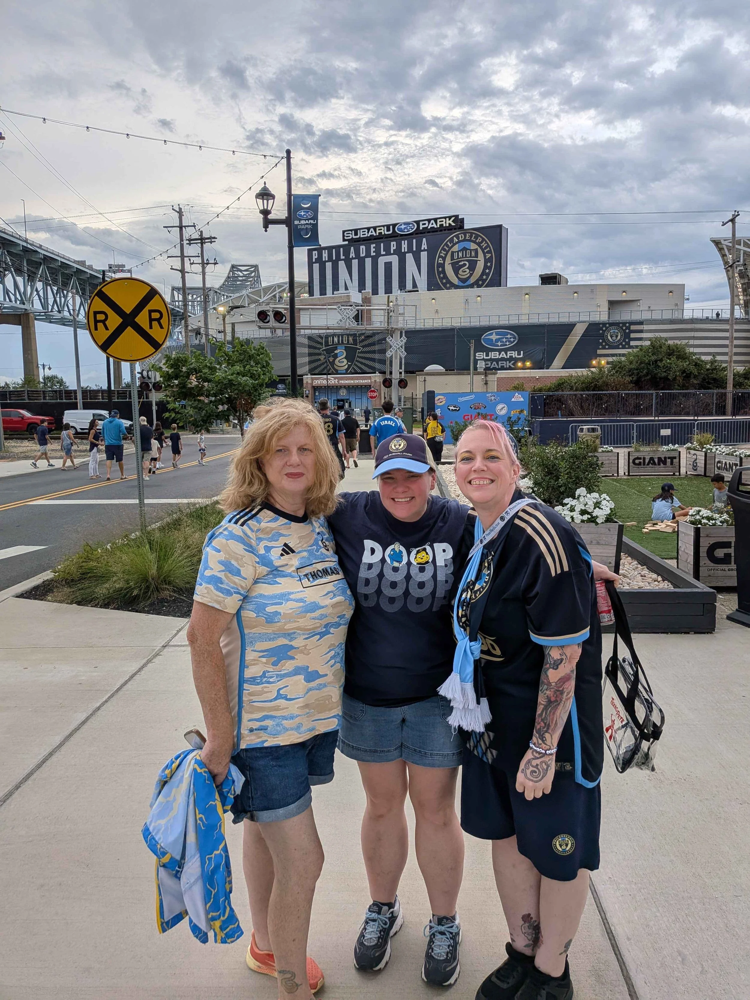 Three women standing together outside at a stadium entrance with a sign that reads 'Philadelphia Union', some people walking in the background, cloudy sky.