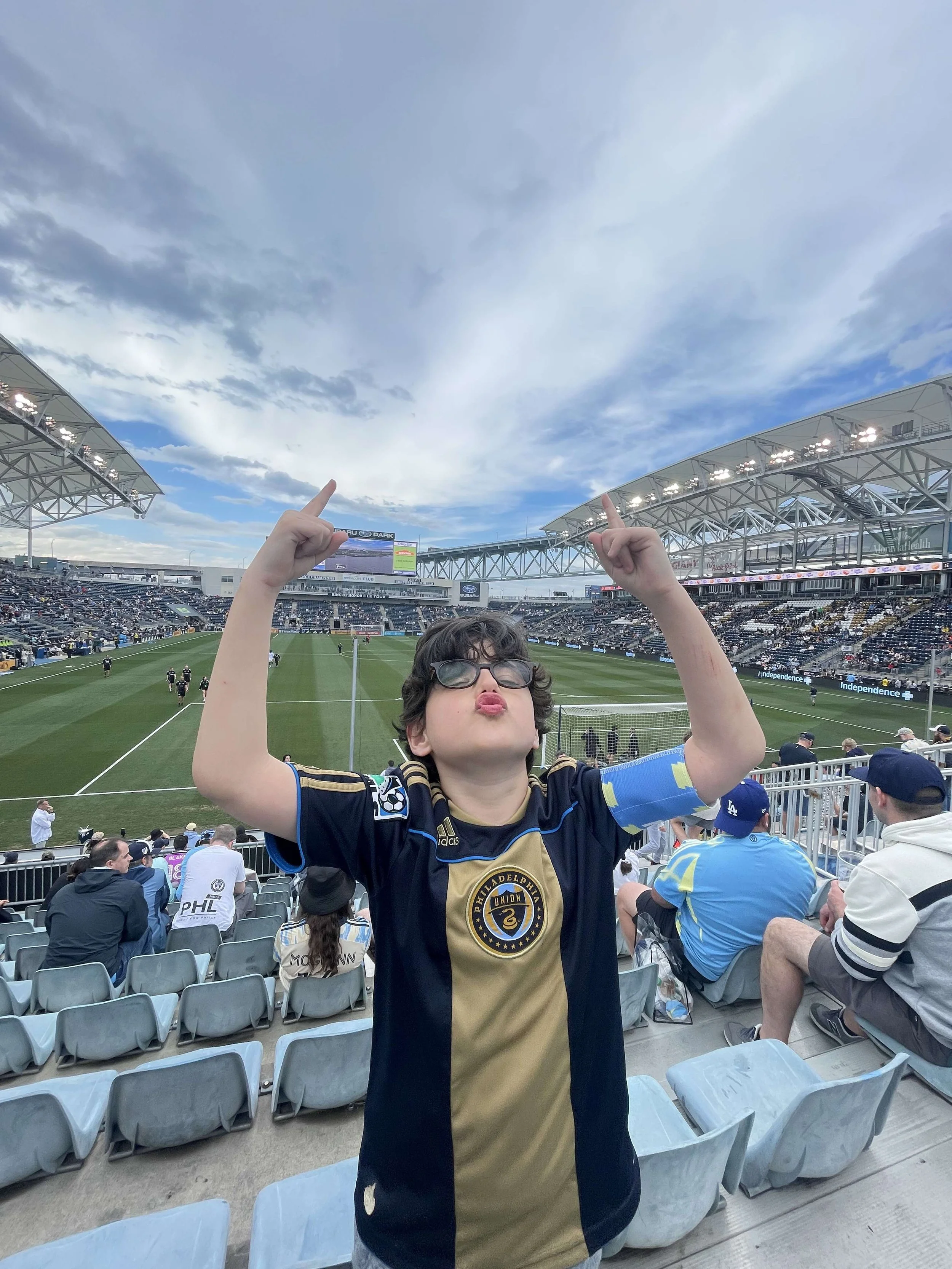 A young boy wearing sunglasses and a Philadelphia Union soccer jersey, making a rock on gesture with both hands at a soccer stadium during a game.