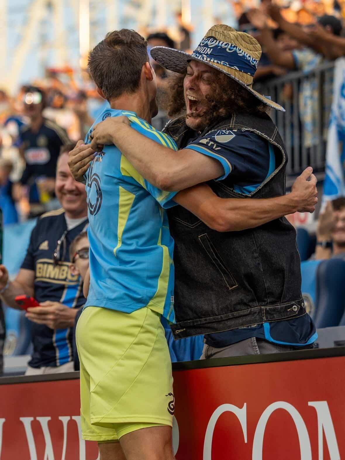 Two men are hugging and celebrating at a sports event, with one in a yellow and blue sports uniform and the other in a black jacket and a large straw hat.