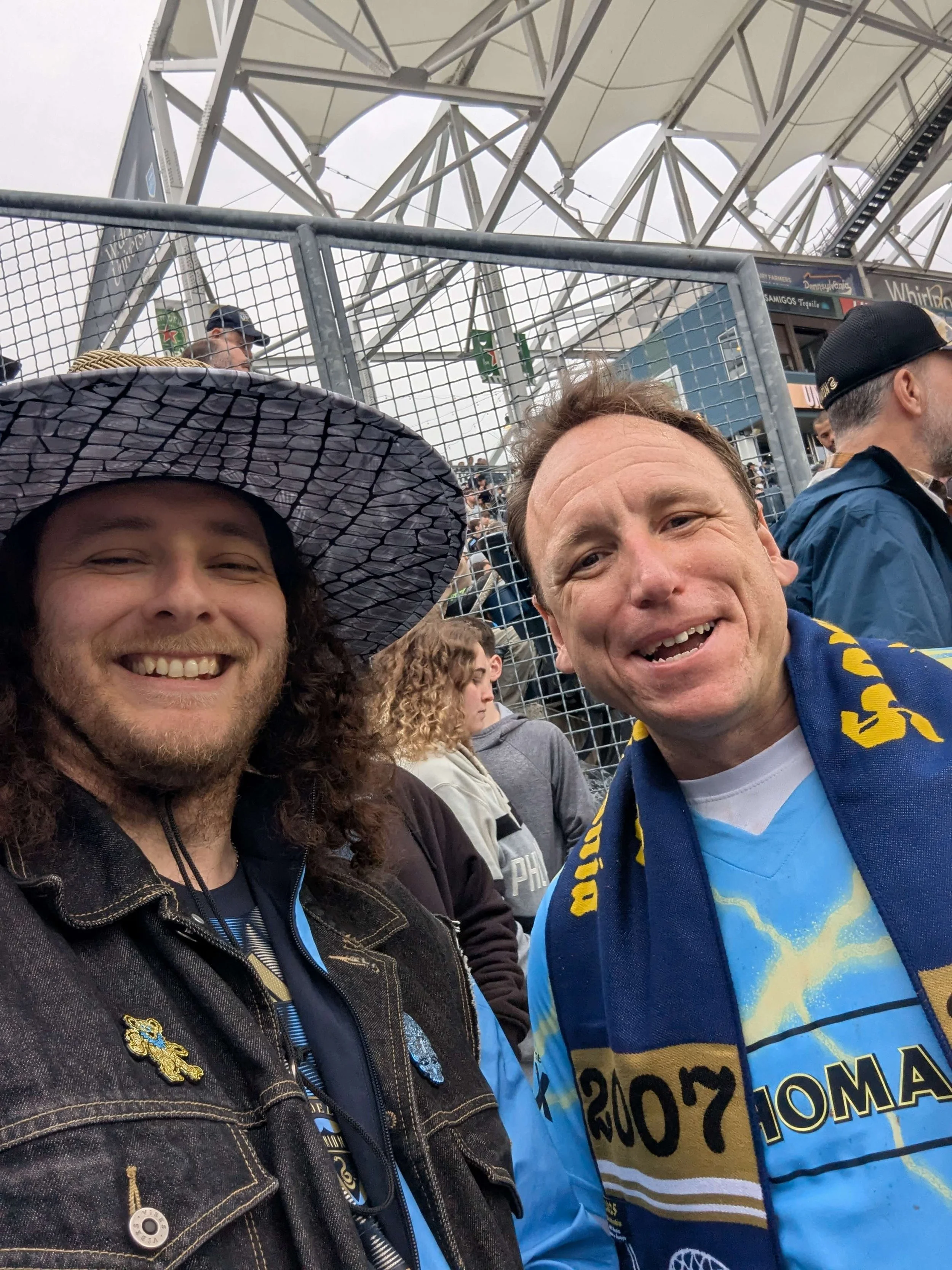 Two smiling men taking a selfie at a crowded outdoor event with a metal fence and stadium seating in the background. One man wears a large hat, the other a blue and yellow scarf. Overcast sky.