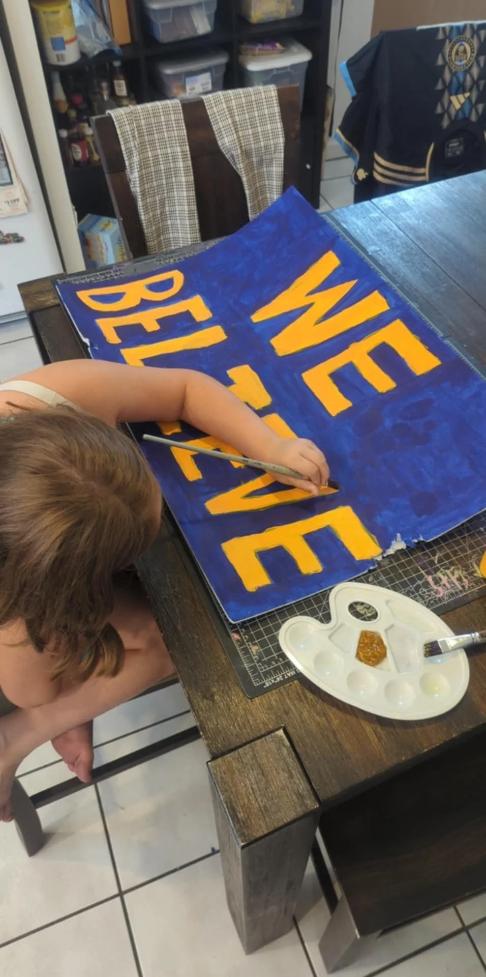 A child painting a sign with yellow letters on a blue background. The sign says 'WED' and 'BELIEVE' and is on a table with a palette and brush. A chair and a cabinet with storage are in the background.