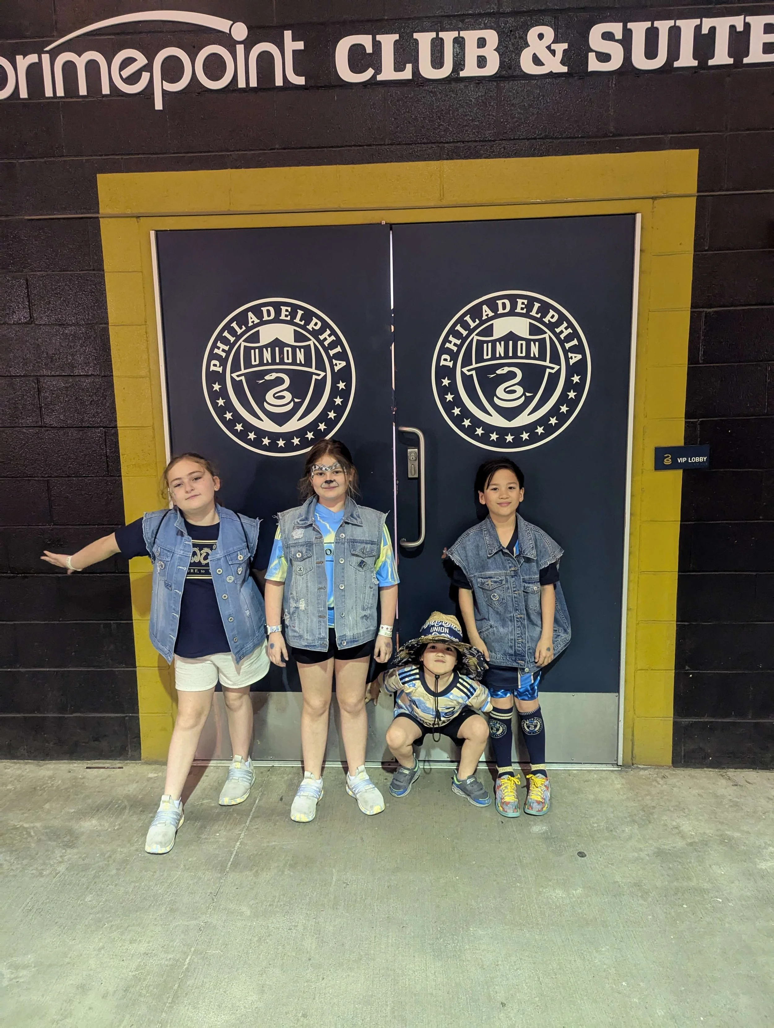 Four children dressed in denim and sports jerseys pose in front of a door featuring the Philadelphia Union logo, inside a club and suite area at the Primepoint stadium.
