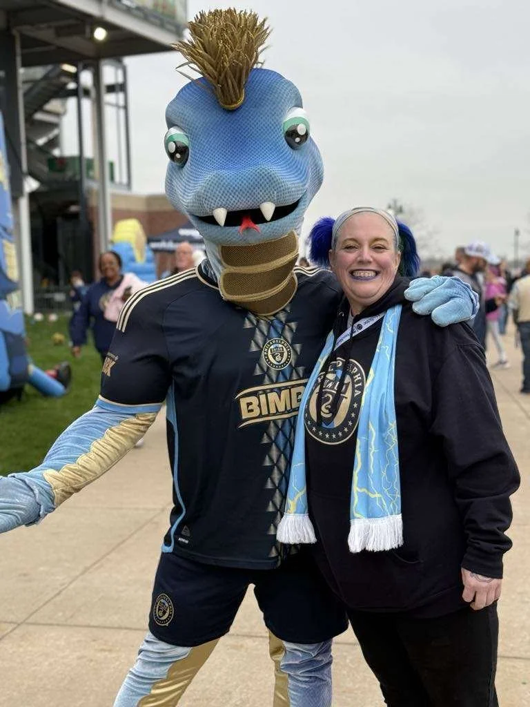 Person dressed in a blue and black soccer jersey and a mascot costume with a dragon head, blue skin, and a mottled brown mane, at an outdoor event with people in the background.