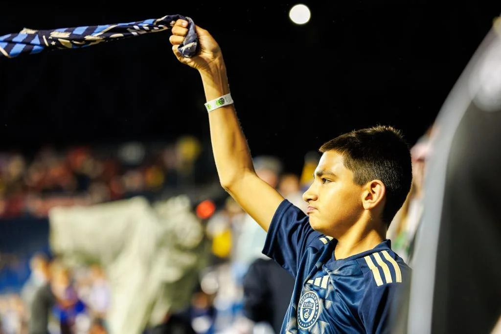 A young boy wearing a blue sports jersey holding a scarf or flag above his head at a sports event or stadium.