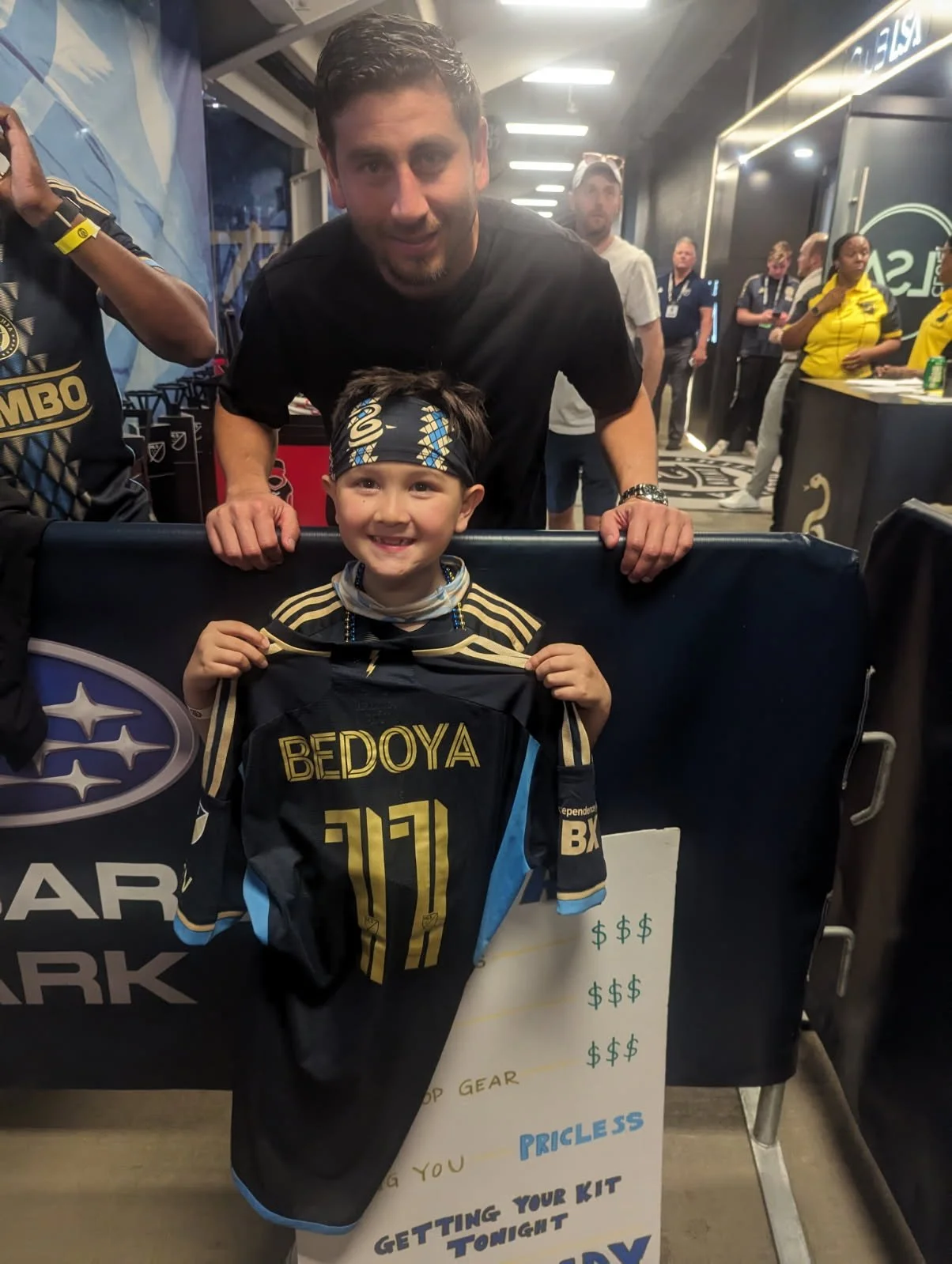 A young boy smiling and holding a black and blue sports jersey with the name 'BEDOYA' and the number 10 on it, standing in front of a table with a sign, while an adult man leans over him in a sports arena or event setting with other people and staff 