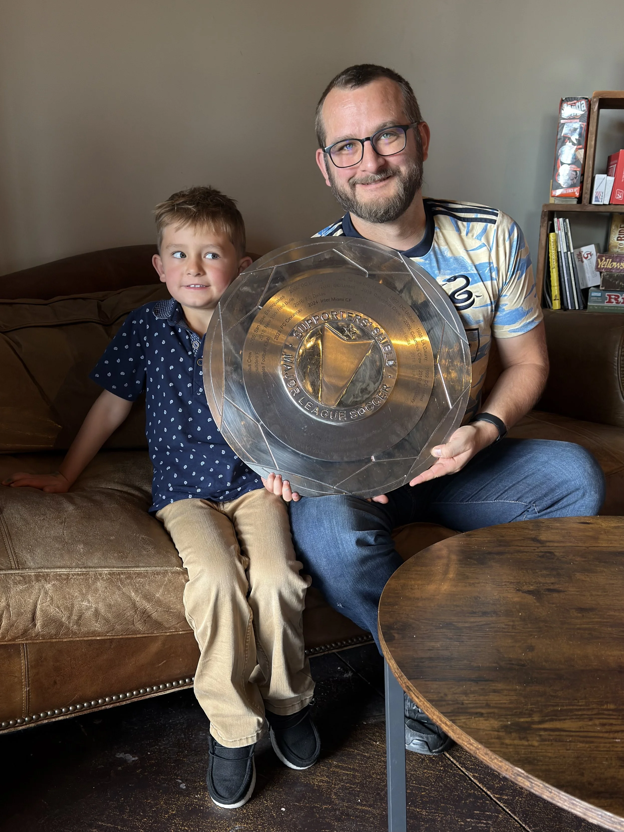A man and his young son sitting on a brown leather couch, holding a clear glass trophy with the words 'Major League Soccer' engraved on it, smiling at the camera.
