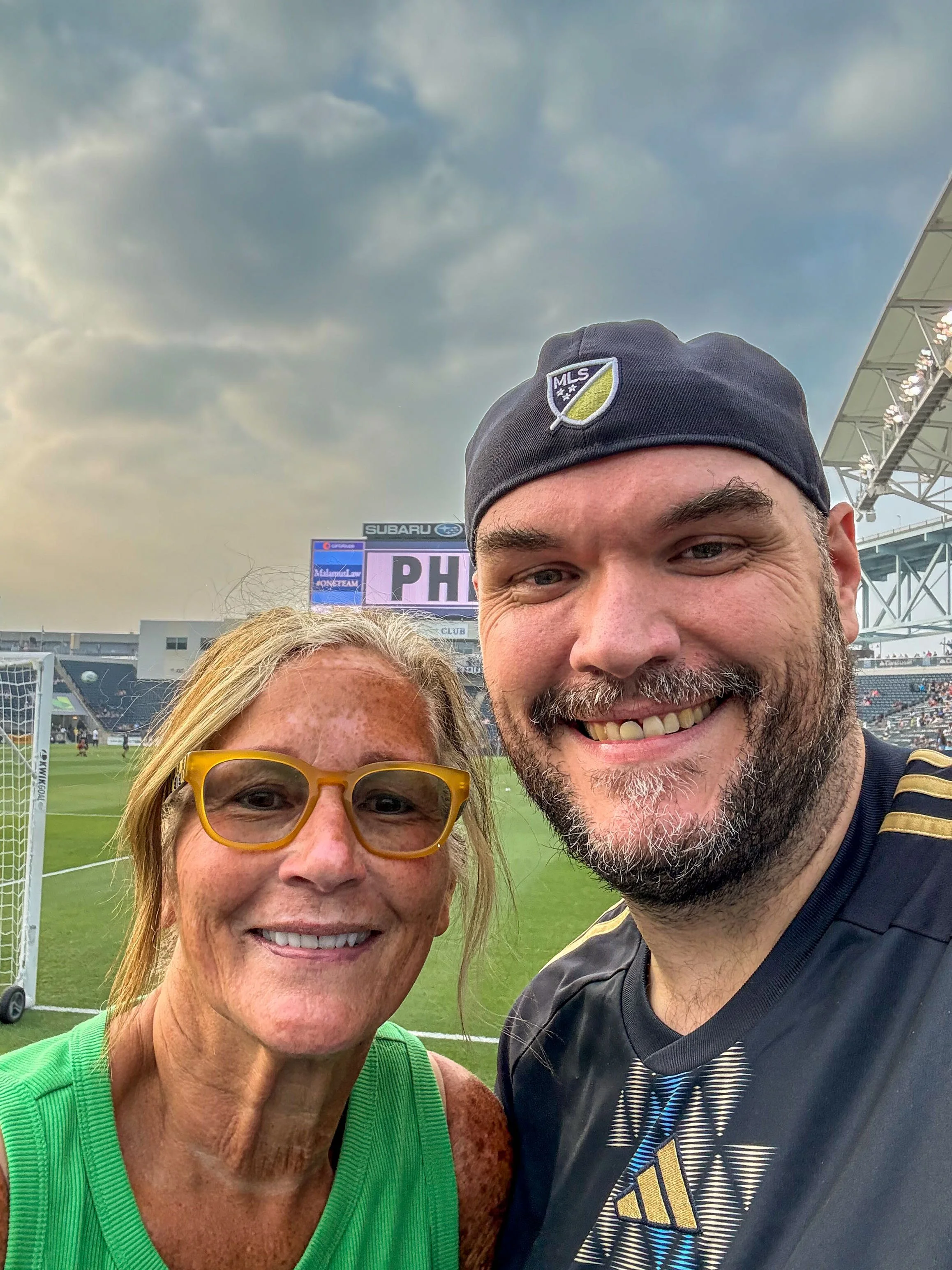 A woman with yellow glasses and a man wearing a black MLS cap taking a selfie at a soccer stadium with cloudy sky.