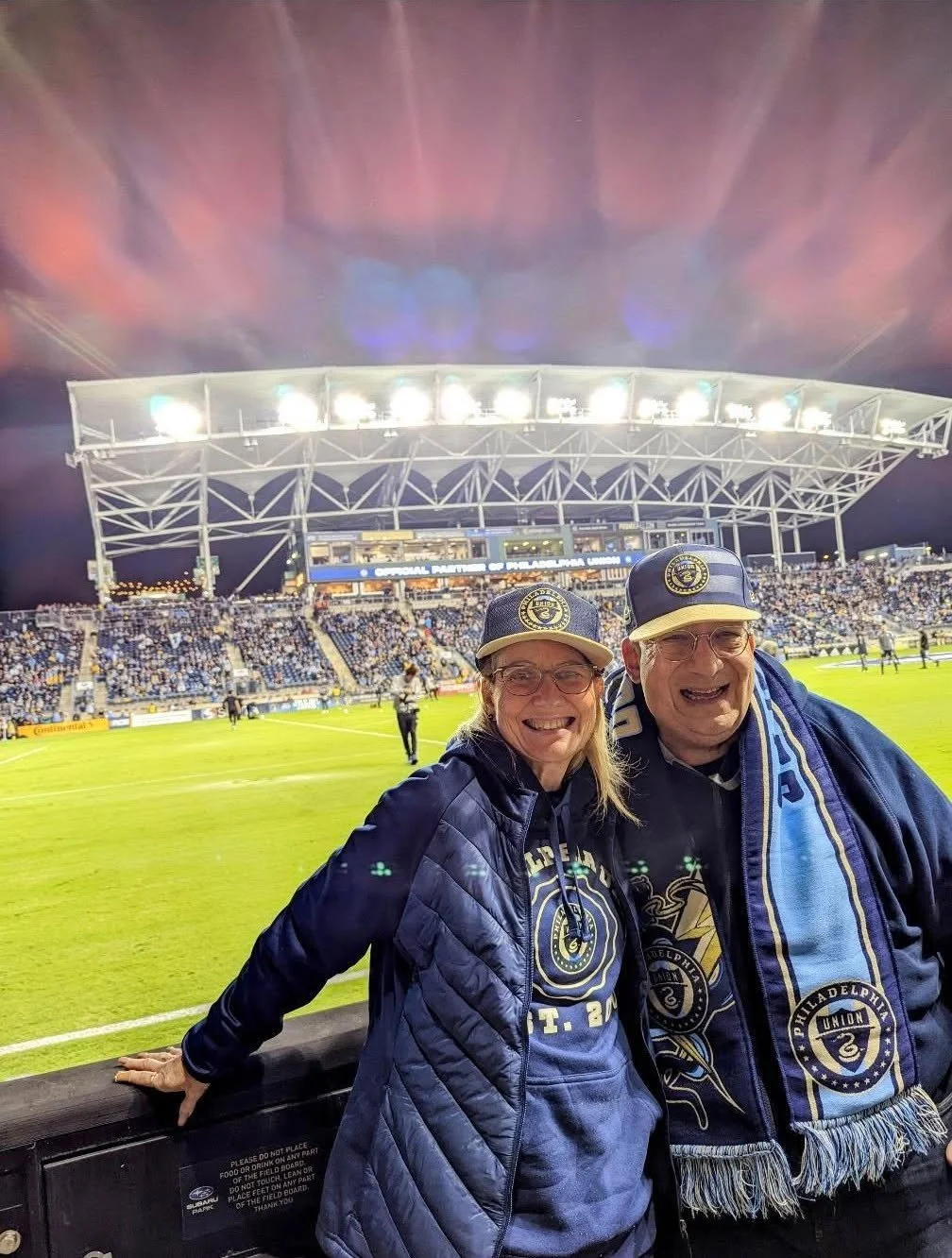 Two adults in Philadelphia Union gear smiling at the camera at a soccer stadium during a night game.