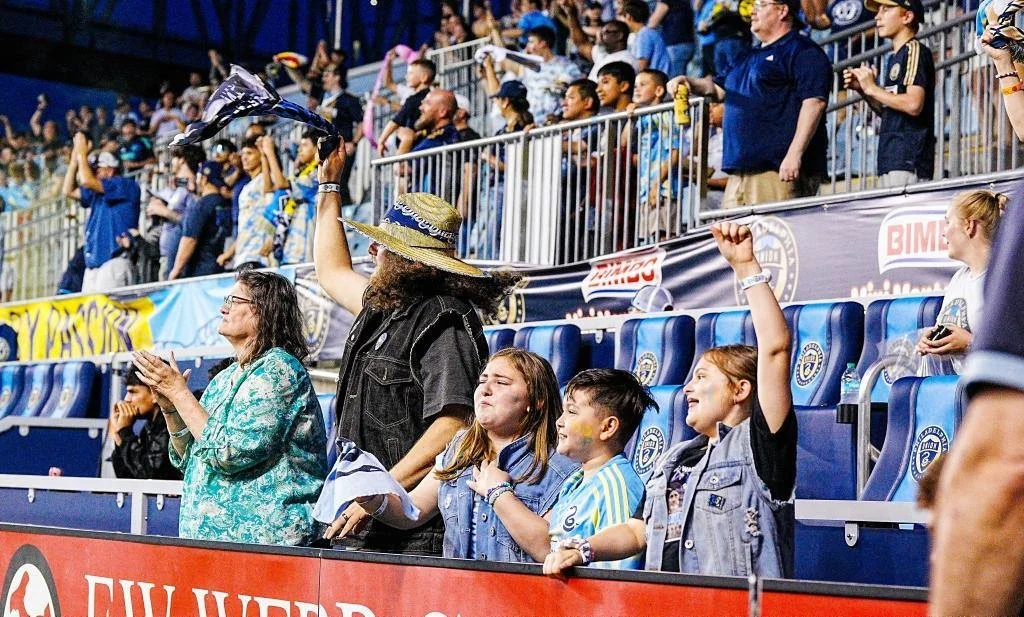 A group of excited soccer fans in the stands, some clapping and cheering, with a woman in a turquoise blouse and a man with a cowboy hat and black denim vest in the foreground. Others are seated or standing, watching the match intently.