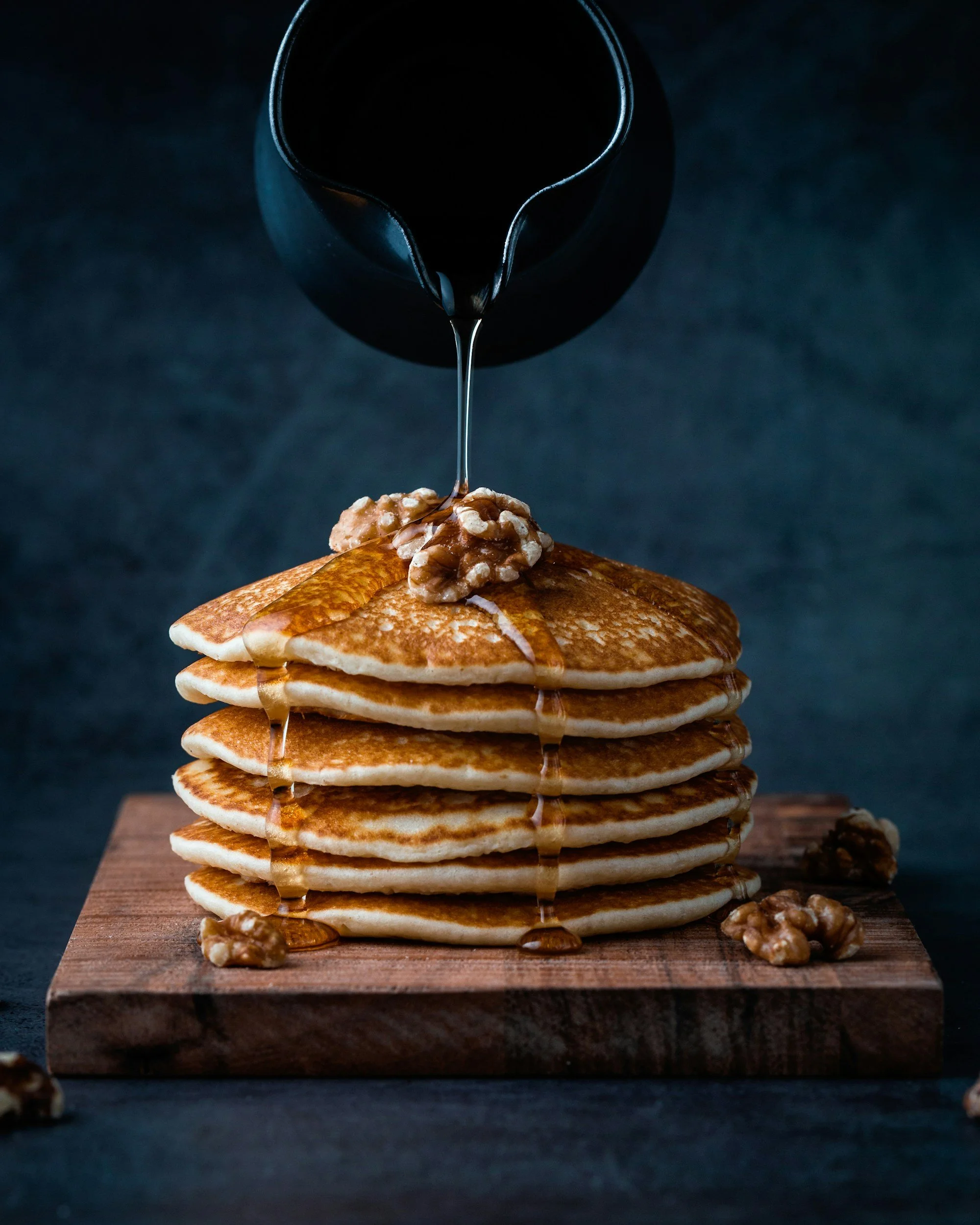 Stack of seven golden-brown pancakes topped with walnuts, with syrup and coffee being poured over them on a wooden cutting board.