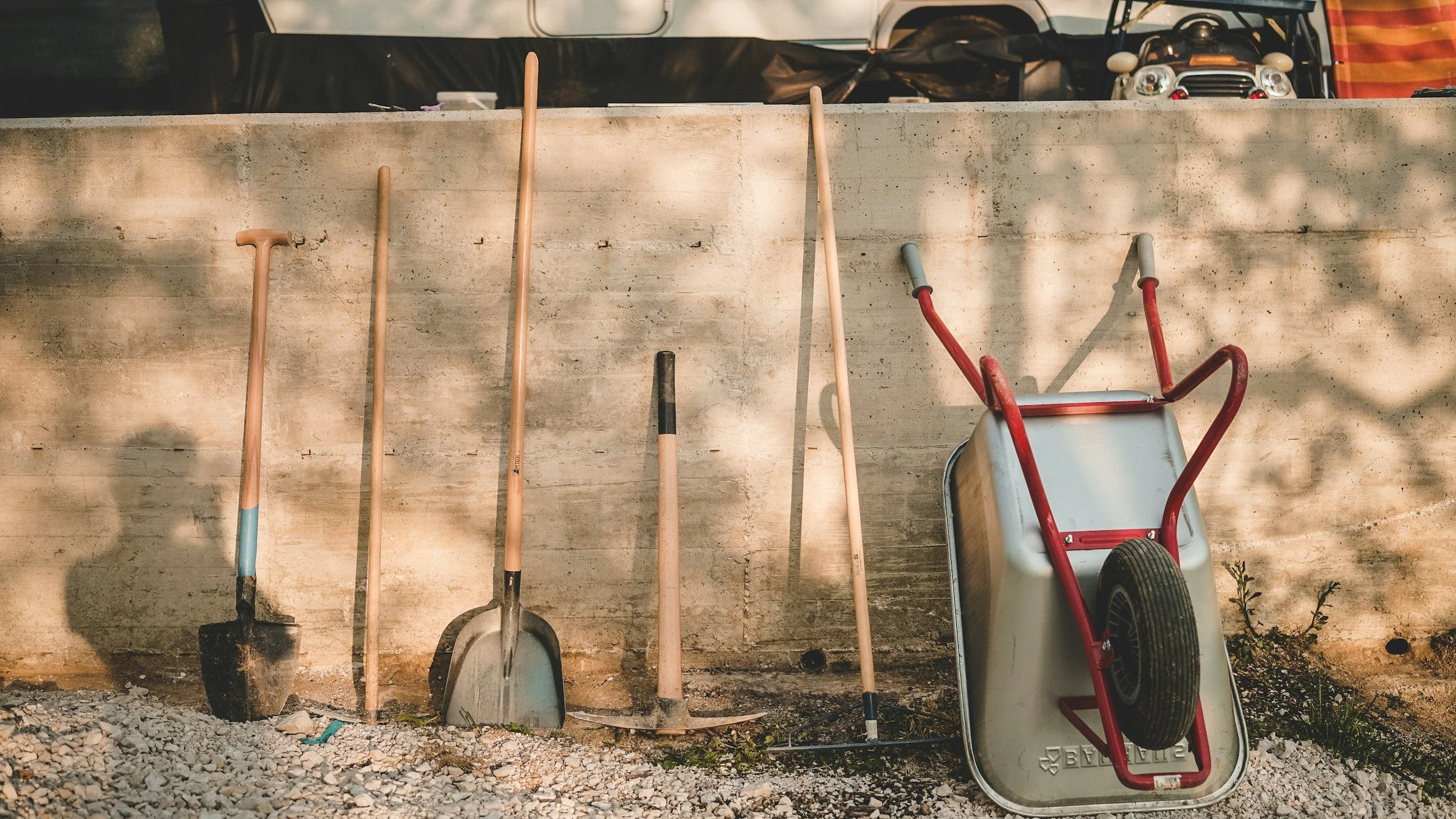 Five gardening tools, including a shovel and a spade, are leaning against a concrete wall. There is a wheelbarrow resting on its side next to the tools, and part of a toy car is visible in the background.