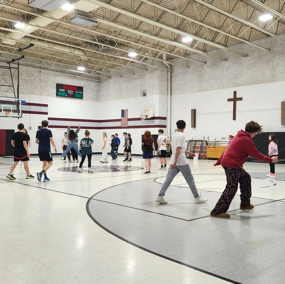 Students playing basketball in a school gymnasium with white walls, maroon stripes, and a scoreboard overhead.