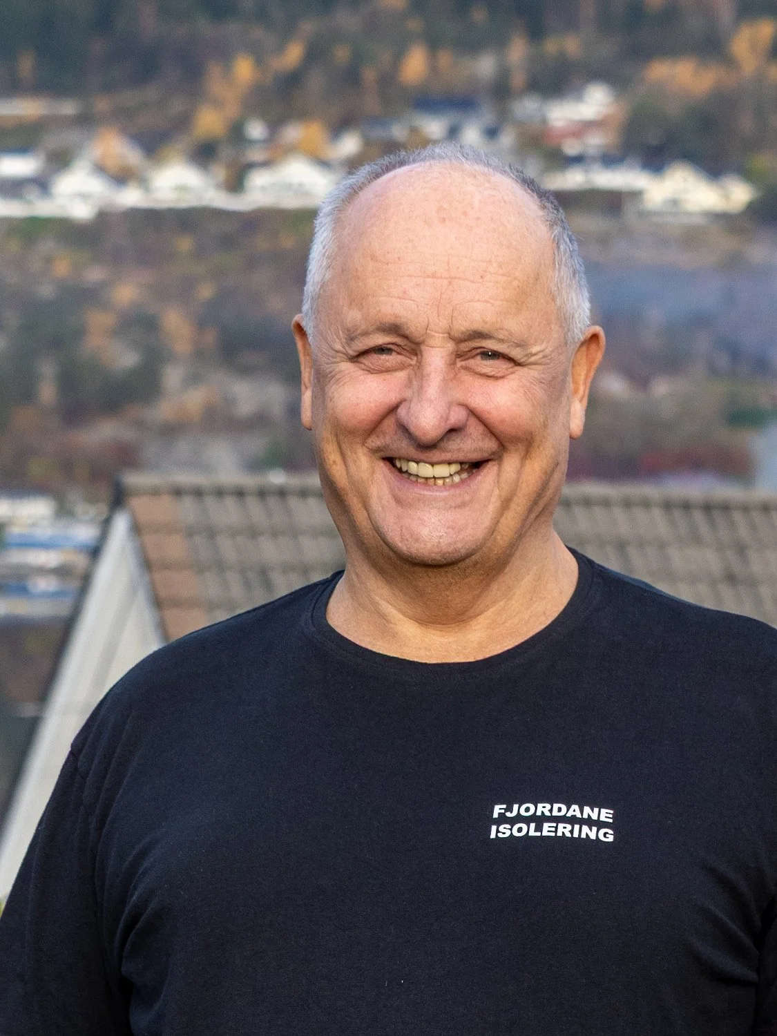 Gunnar Kjørvik er ein triveleg og arbeidssam kar, her iført svart arbeidsantrekk med t-skjorte med teksten 'FJORDANE ISOLERING'. Han står utendørs med en landsby og fjell i bakgrunnen.