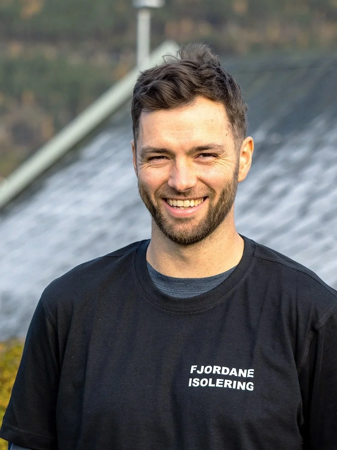 En mann smiler og står foran en vannkraft- eller hydroelektrisk anlegg, iført svart t-skjorte med teksten 'FJORDANE ISOLERING'.