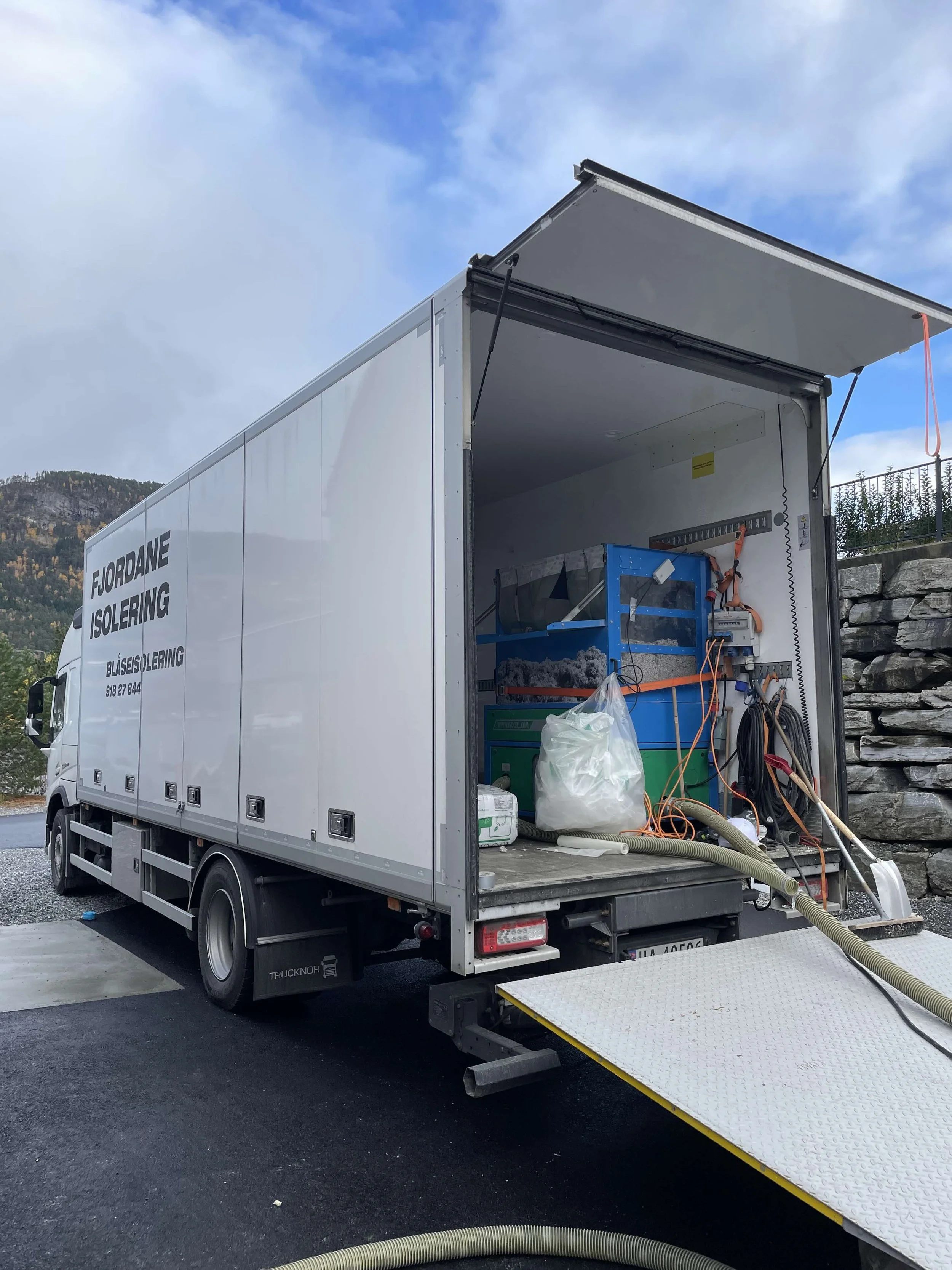 Lastebil med åpen bakdør, utstyr for fjord- og isoleringsarbeid, rør og verktøy inne i lastebilen, med fjell og blå himmel i bakgrunnen.
