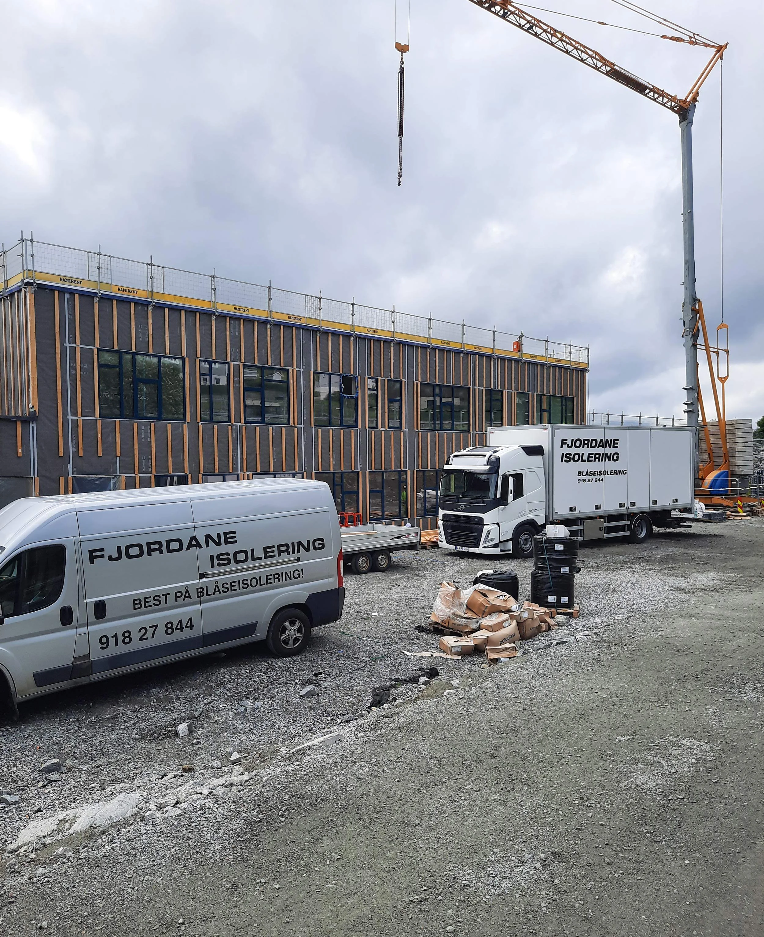 Utsikt av en byggeplass med to varebiler og en lastebil med skiltet Fjordane Isolering, en stor kran, og et bygning under oppføring.