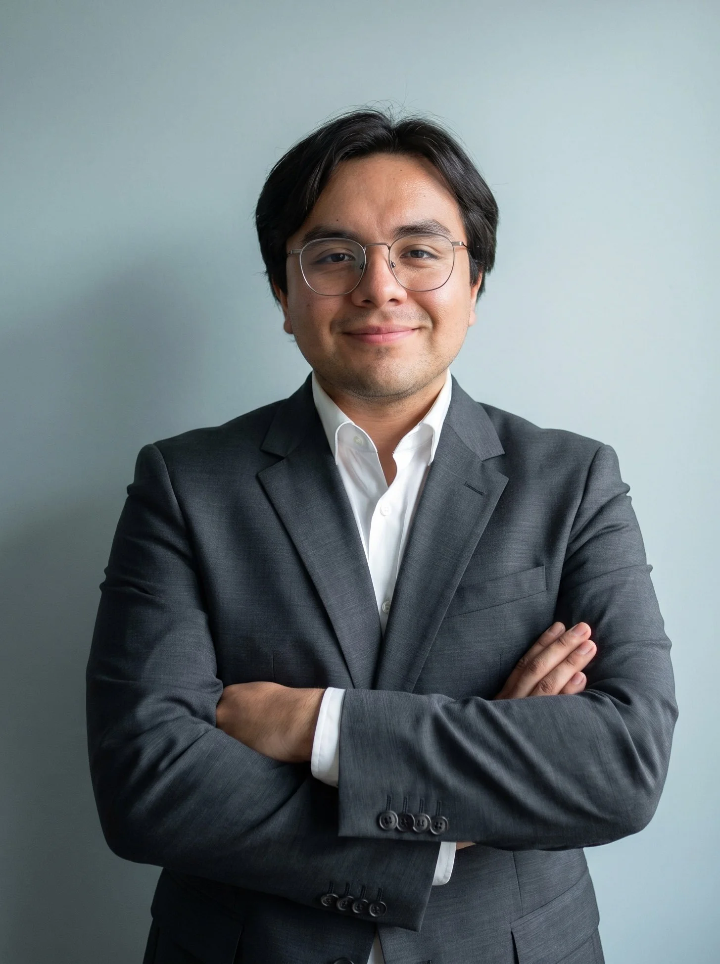 Hombre joven con gafas, vestido con traje gris y camisa blanca, posando con los brazos cruzados y sonriendo, contra un fondo gris claro.