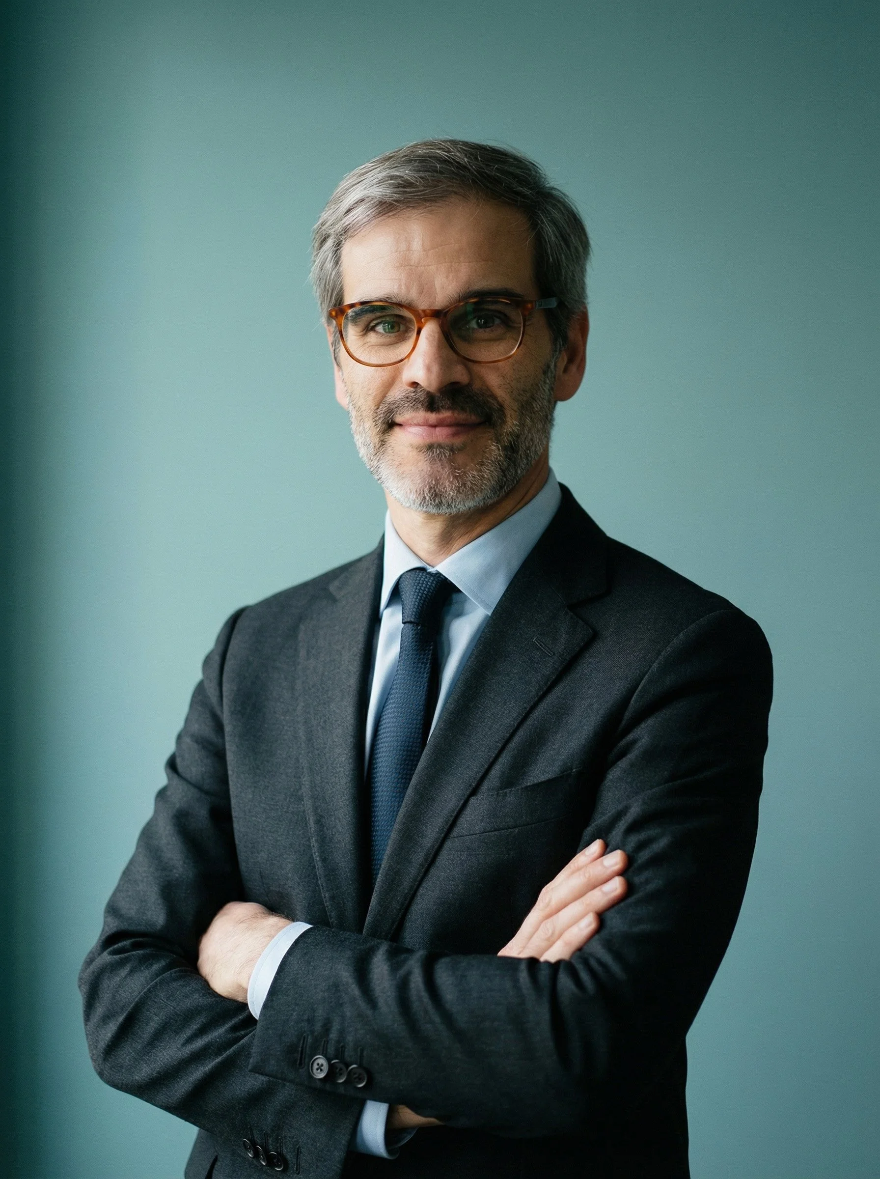 Hombre con gafas de abogado o ejecutivo en traje, con barba y cabello canoso, sonriendo y cruzado de brazos, fondo azul claro.