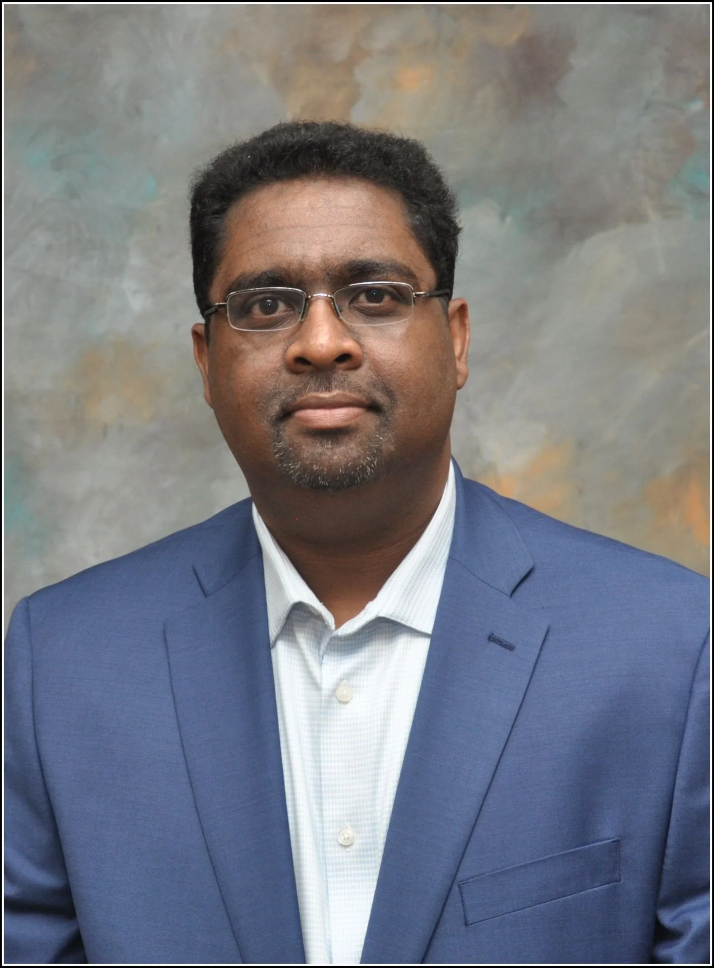 A man wearing glasses and a blue suit jacket with a white collared shirt, posing in front of a textured gray and beige background.