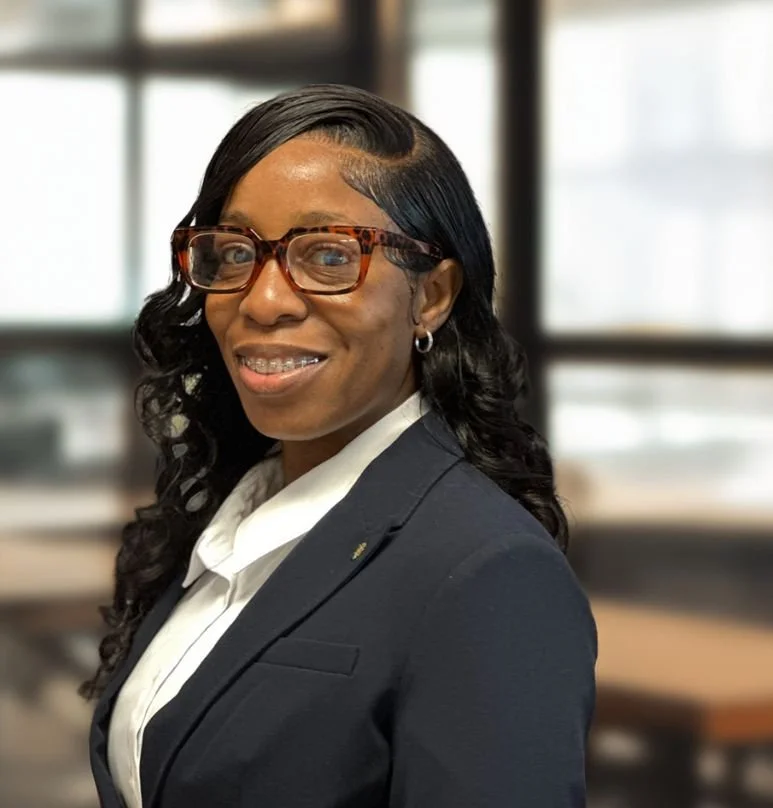 A woman with long dark wavy hair, glasses, and earrings, wearing a dark blazer and white shirt, smiling in an office setting.