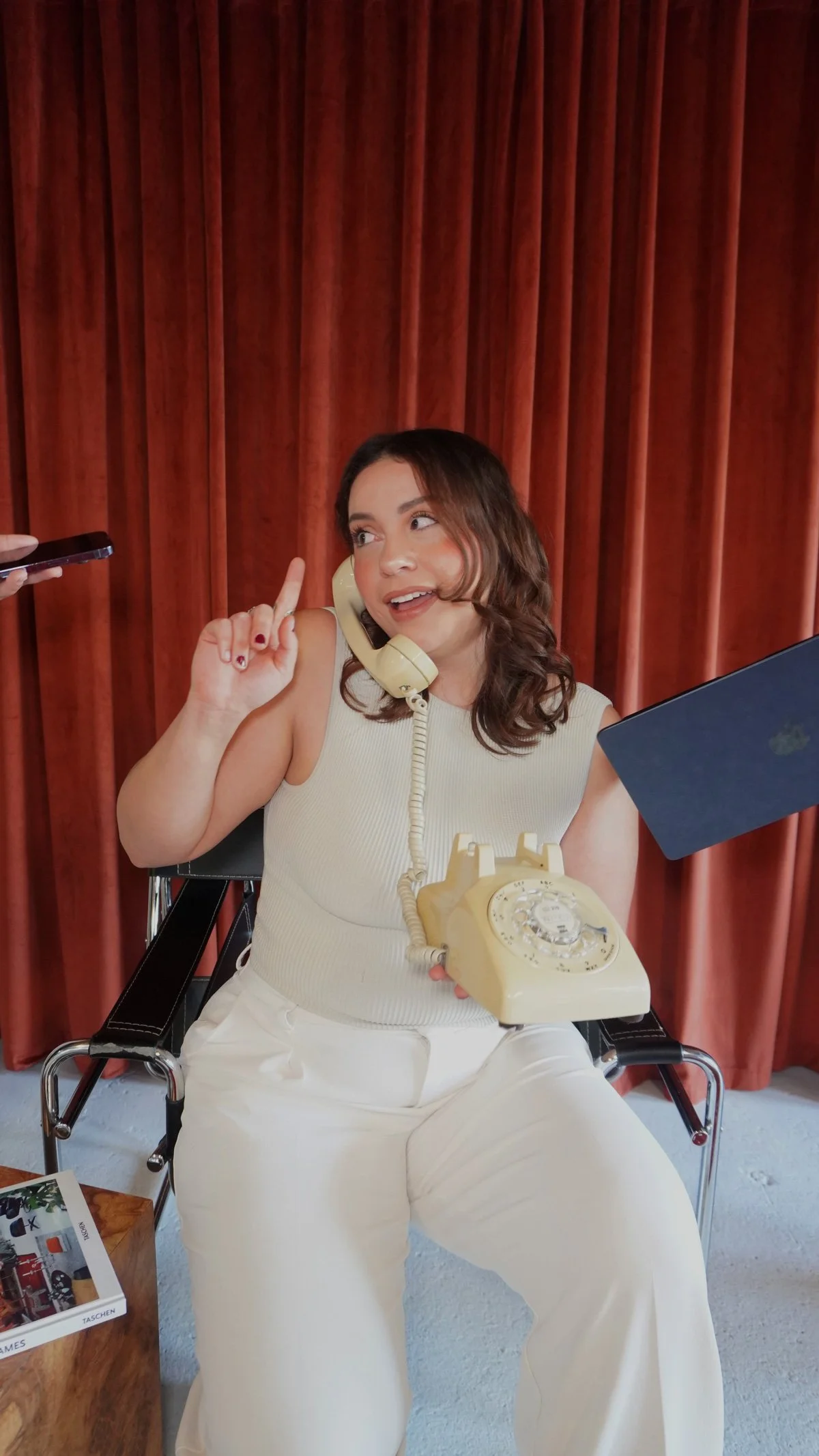 Woman sitting on a chair with a vintage telephone on her lap, holding a blue folder, and speaking on a beige rotary phone with a cord, in front of a red curtain.