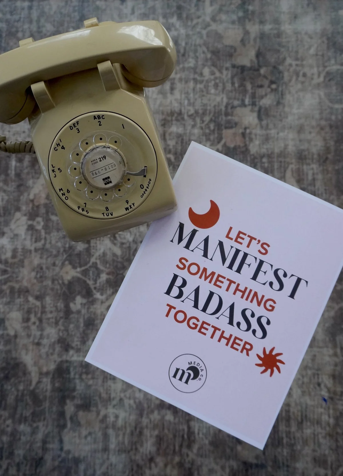 A vintage beige rotary telephone placed on a wooden surface next to a motivational print that reads 'Let's Manfest Something Badass Together' with a small logo at the bottom.