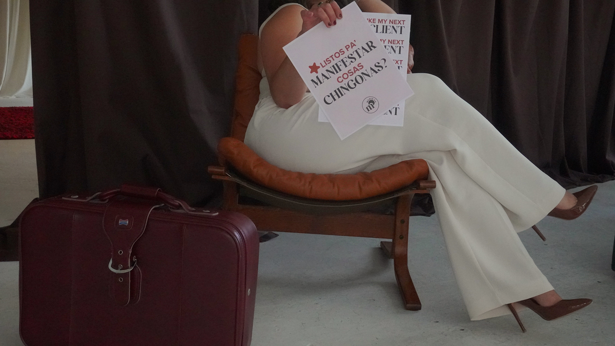 Person sitting on a wooden chair with a brown leather cushion, holding protest signs, with a red suitcase on the floor nearby.